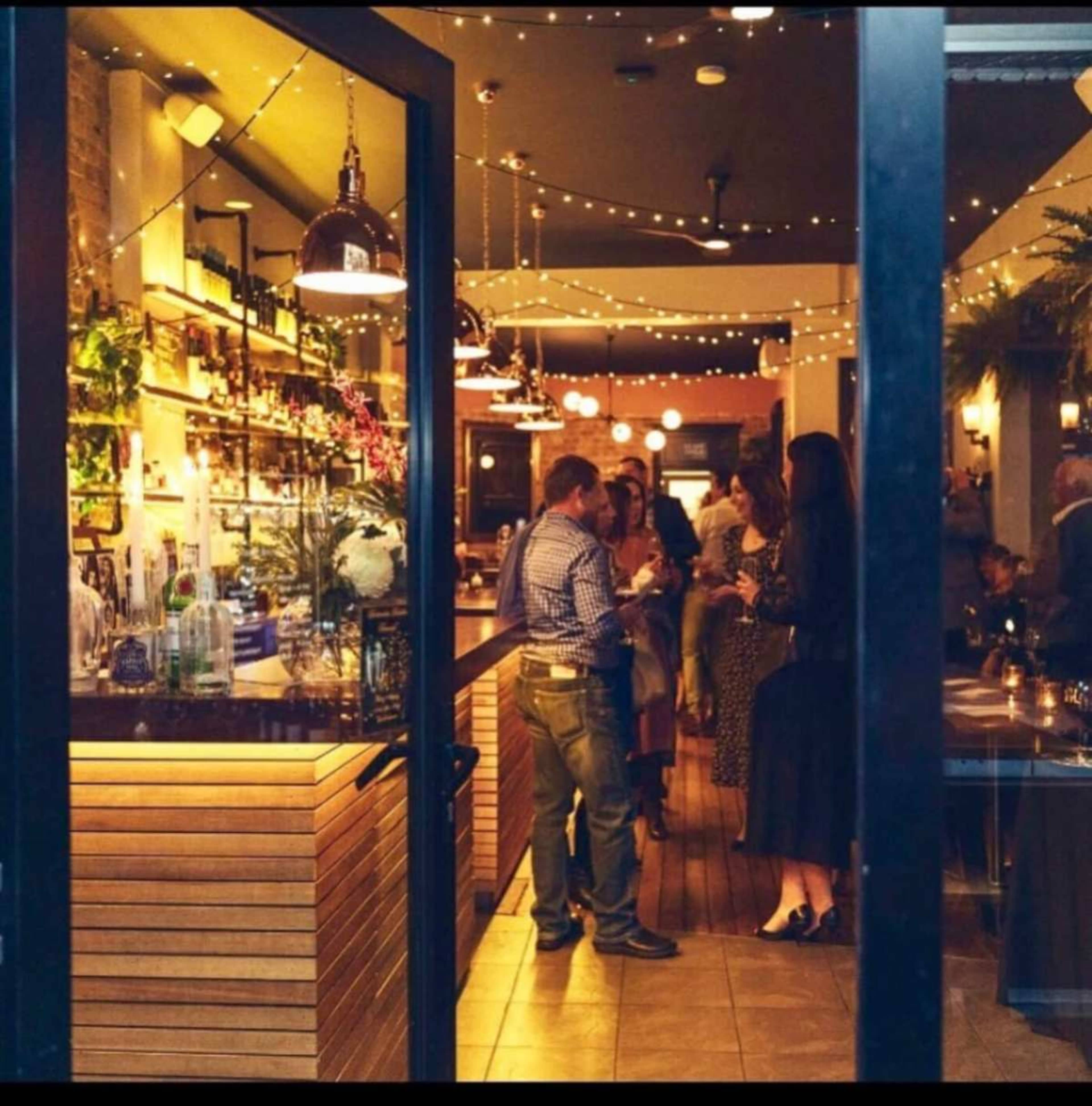 The image shows a well-lit bar area with patrons socializing, surrounded by shelves of liquor and decorative string lights.
