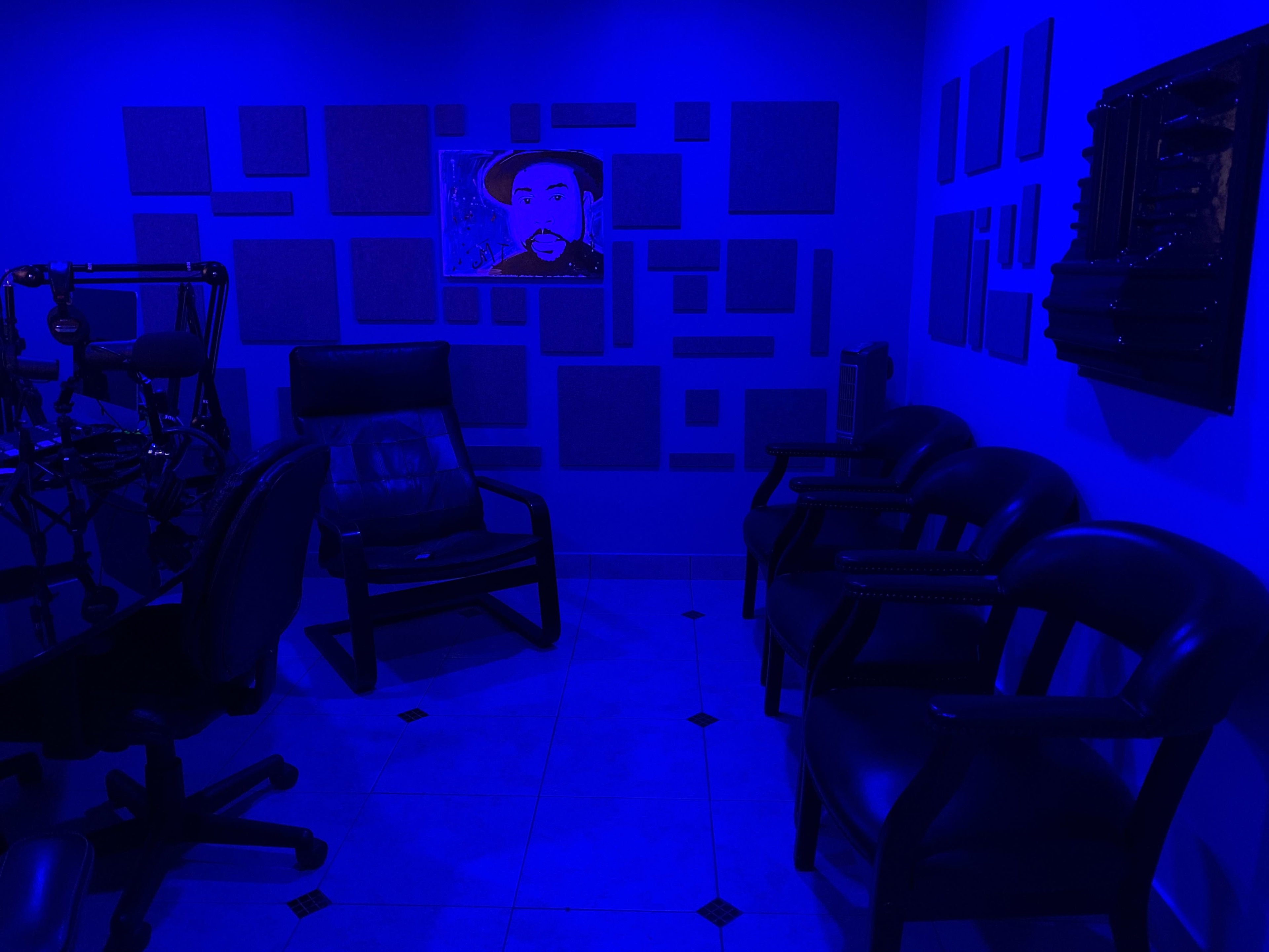 A dimly lit room with blue walls featuring soundproof panels, a painting on the wall, and several black chairs arranged around a table.