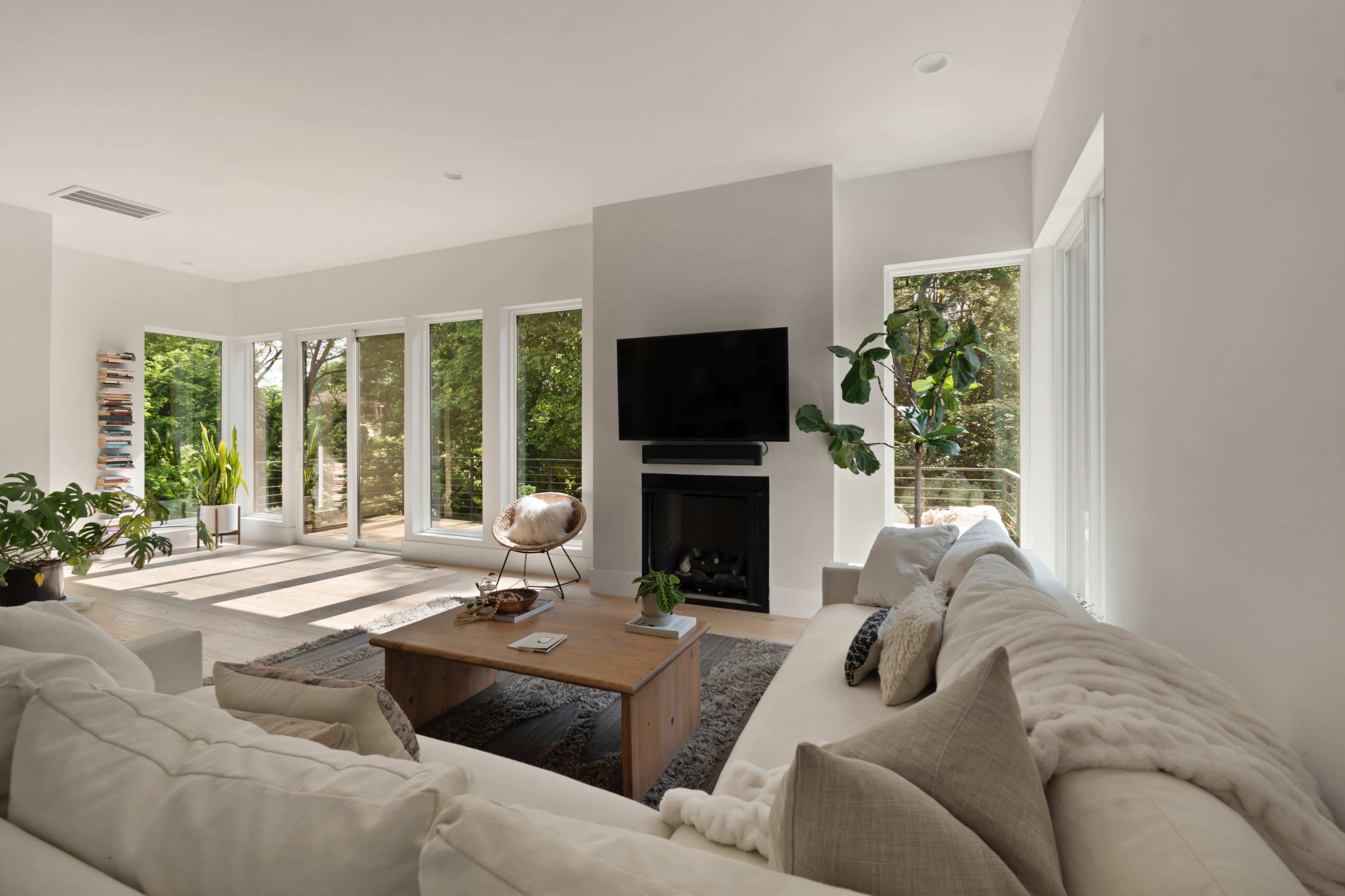 A modern living room features a large sectional sofa, a coffee table, a flat-screen TV mounted above a fireplace, and large windows that provide a view of greenery outside.