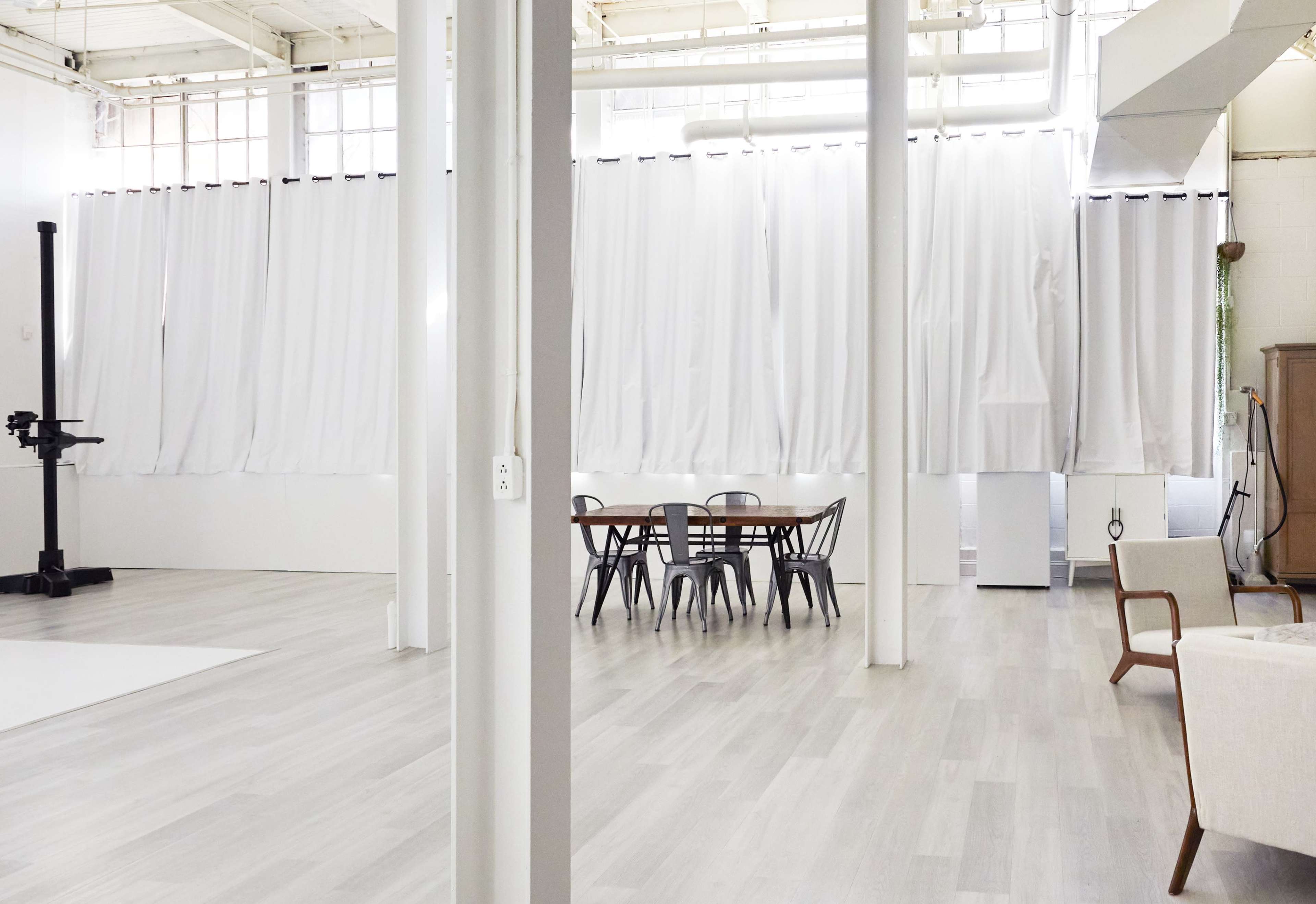 The image shows a spacious, modern interior with white curtains, wooden furniture, and large windows allowing natural light.