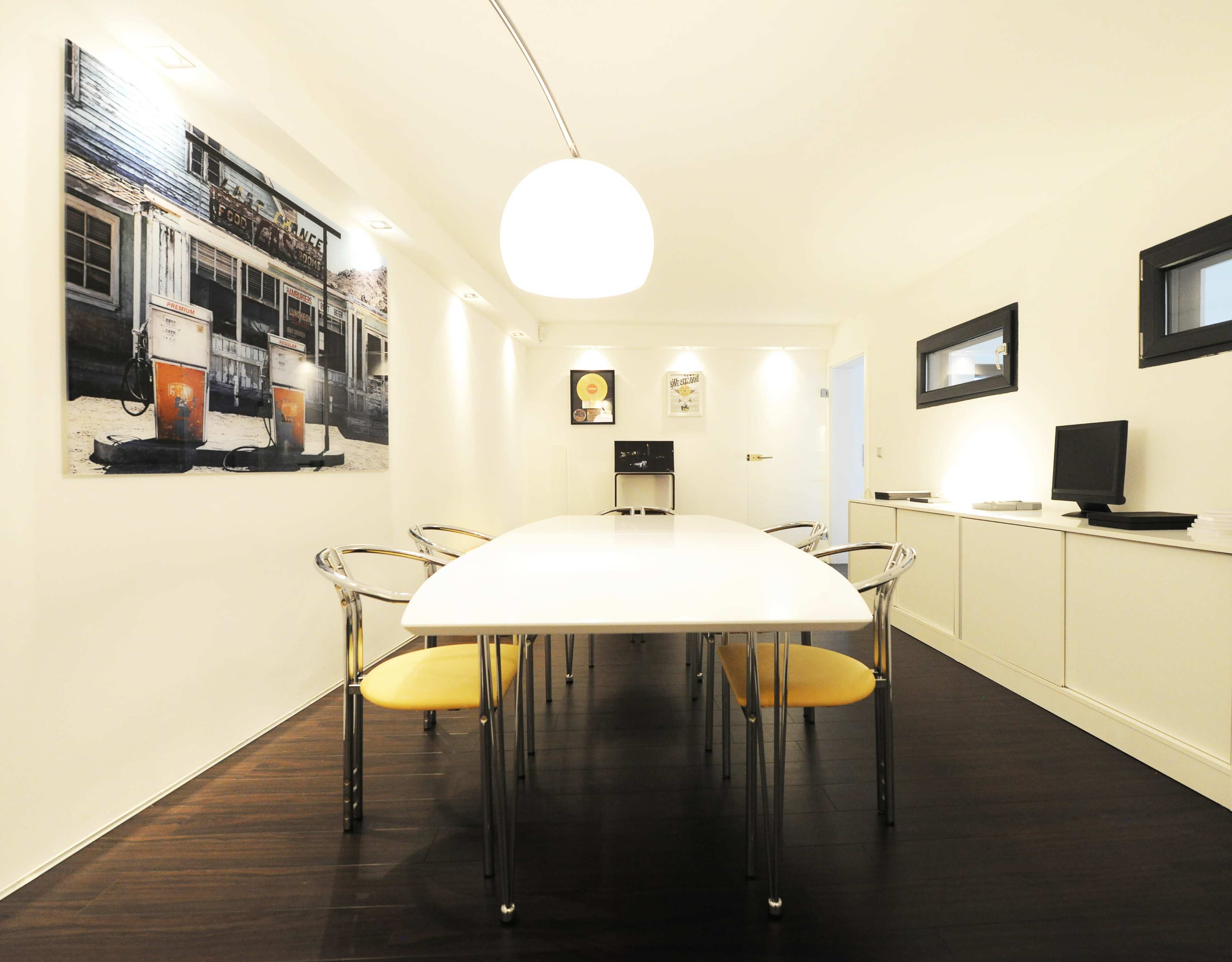 The image shows a modern meeting room with a long white table surrounded by transparent chairs and a large wall-mounted photograph.