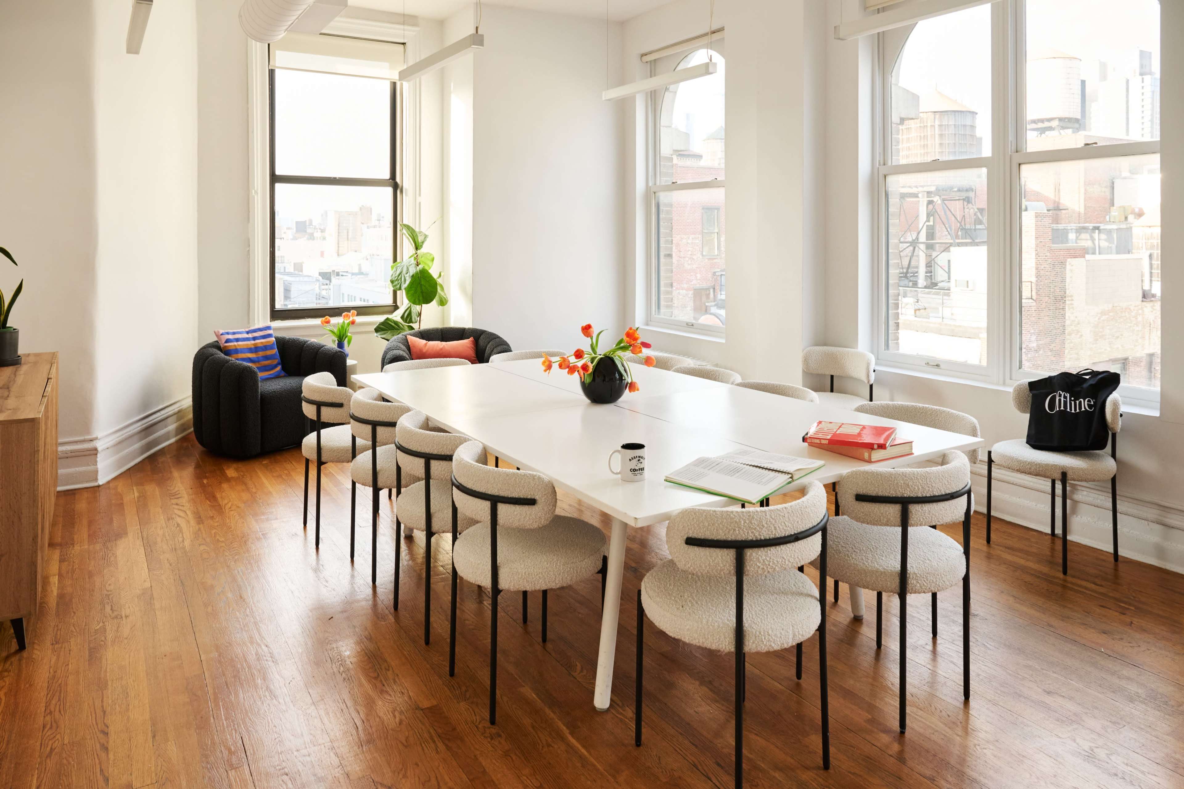A spacious, brightly lit conference room features a long white table surrounded by ten chairs, with a green plant and decorative items on the table and a cozy seating area in the background.
