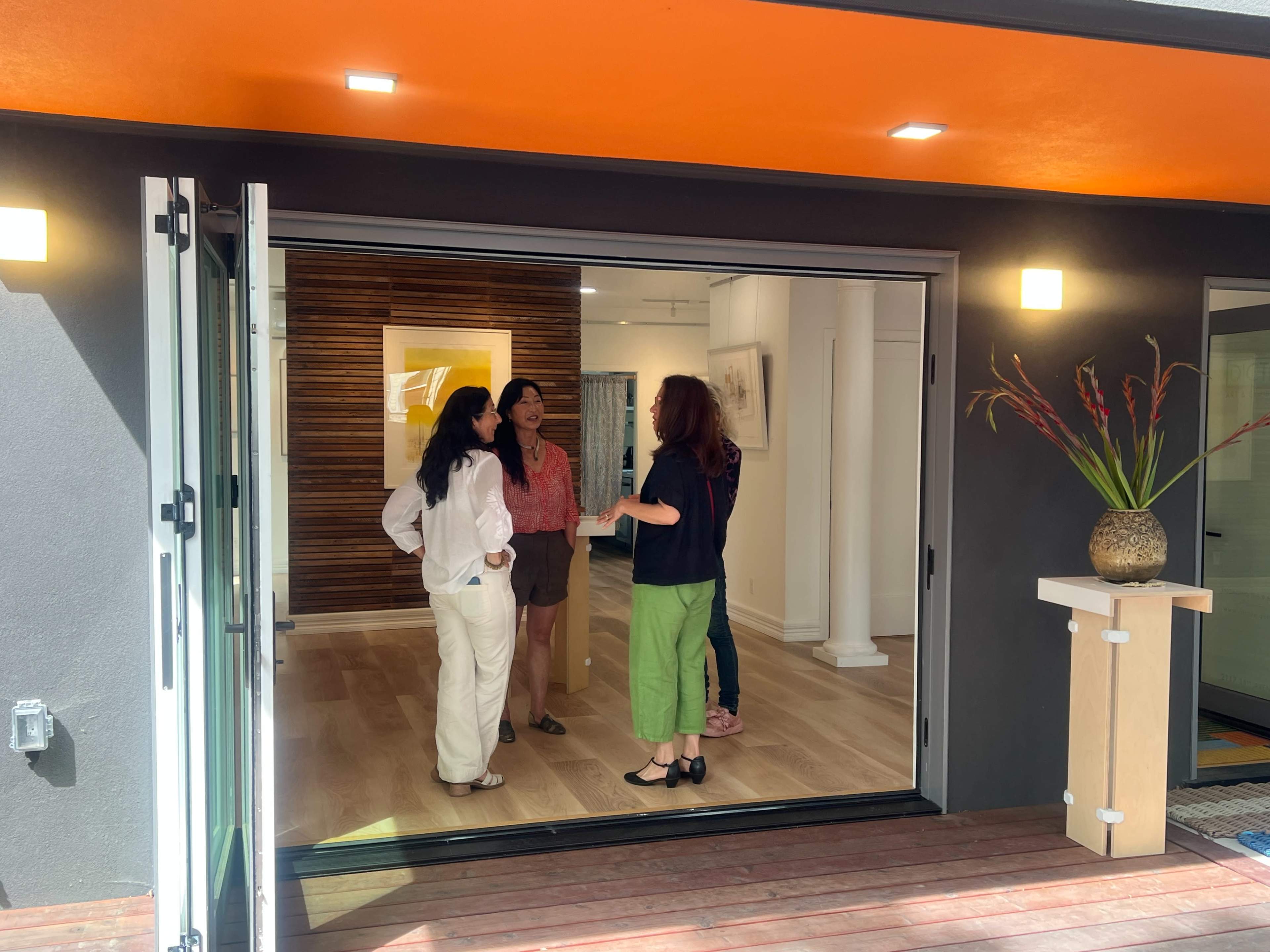 Four women are engaged in conversation inside an art gallery that features wooden floors and an orange ceiling.