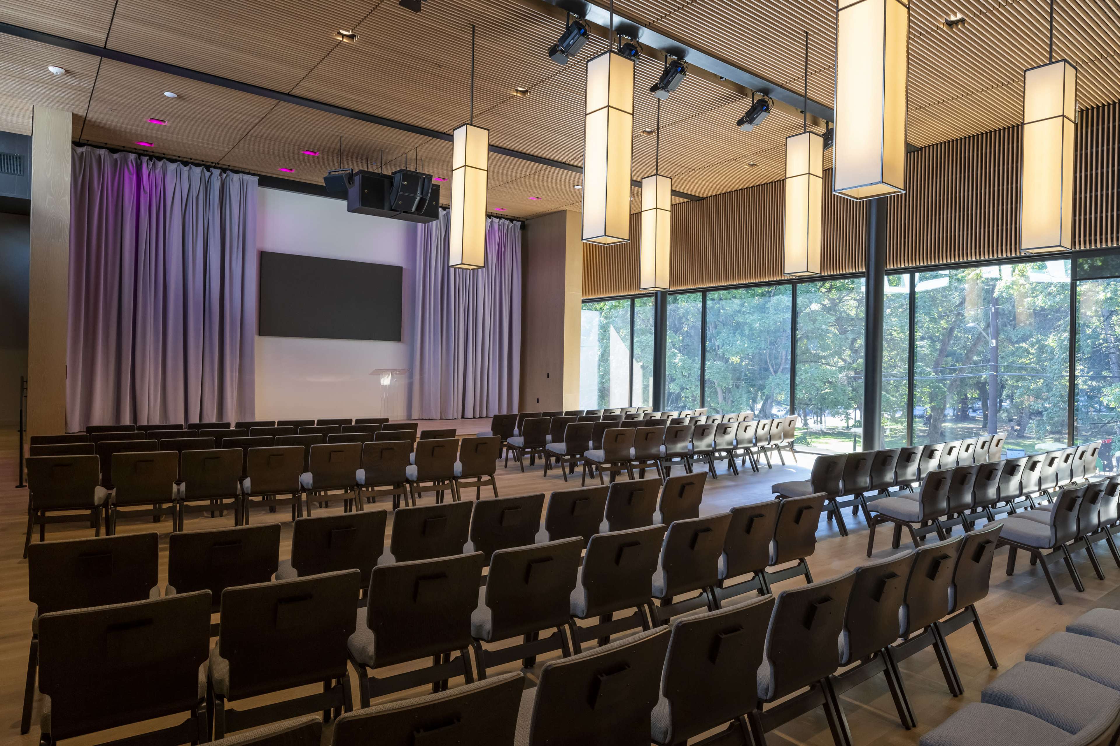 A modern auditorium features rows of chairs facing a stage with a large screen, large windows allowing natural light to flood the space.