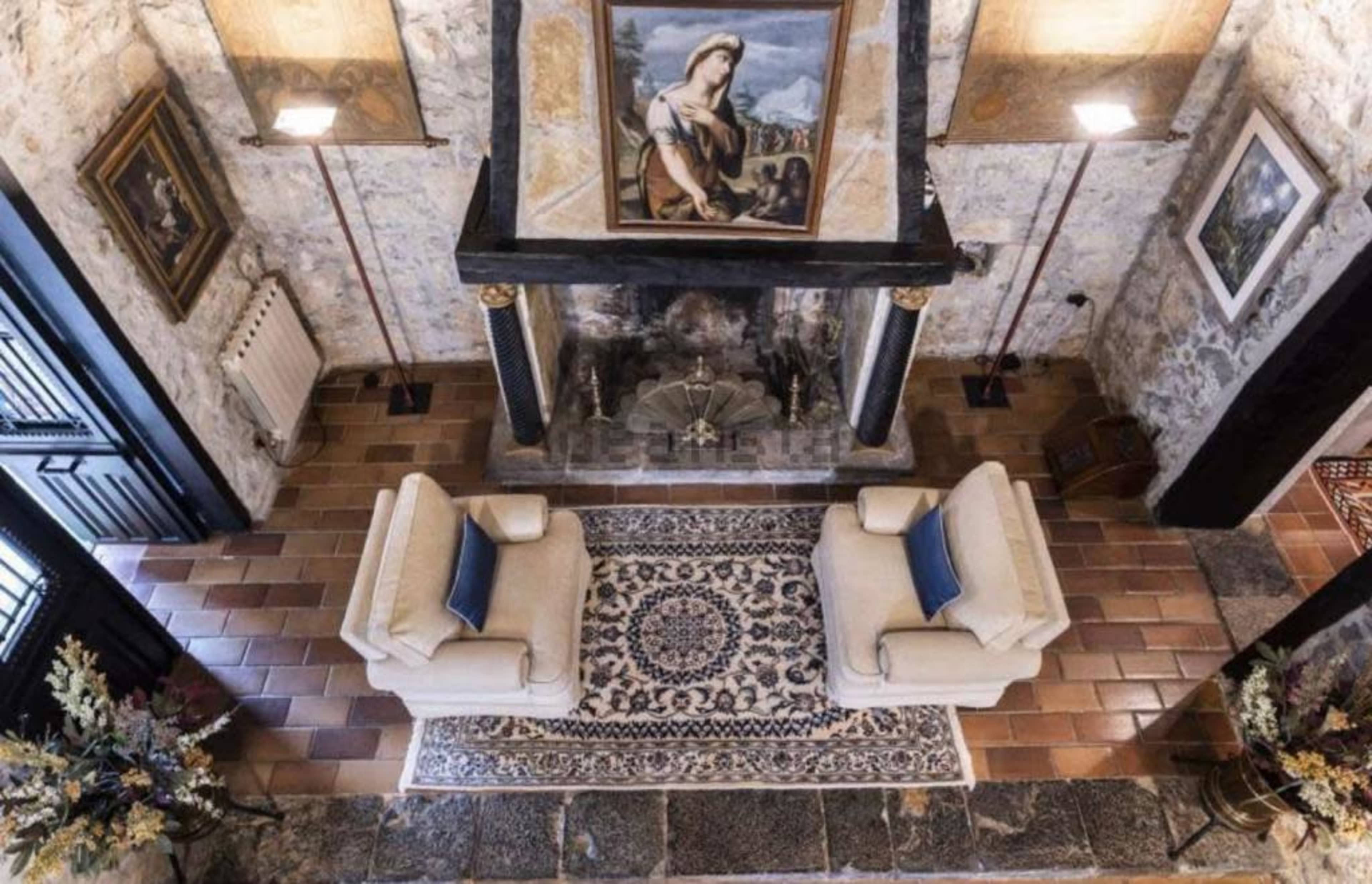 The image shows a cozy living room featuring two armchairs facing a stone fireplace, with a painting above the mantel and patterned tiles on the floor.