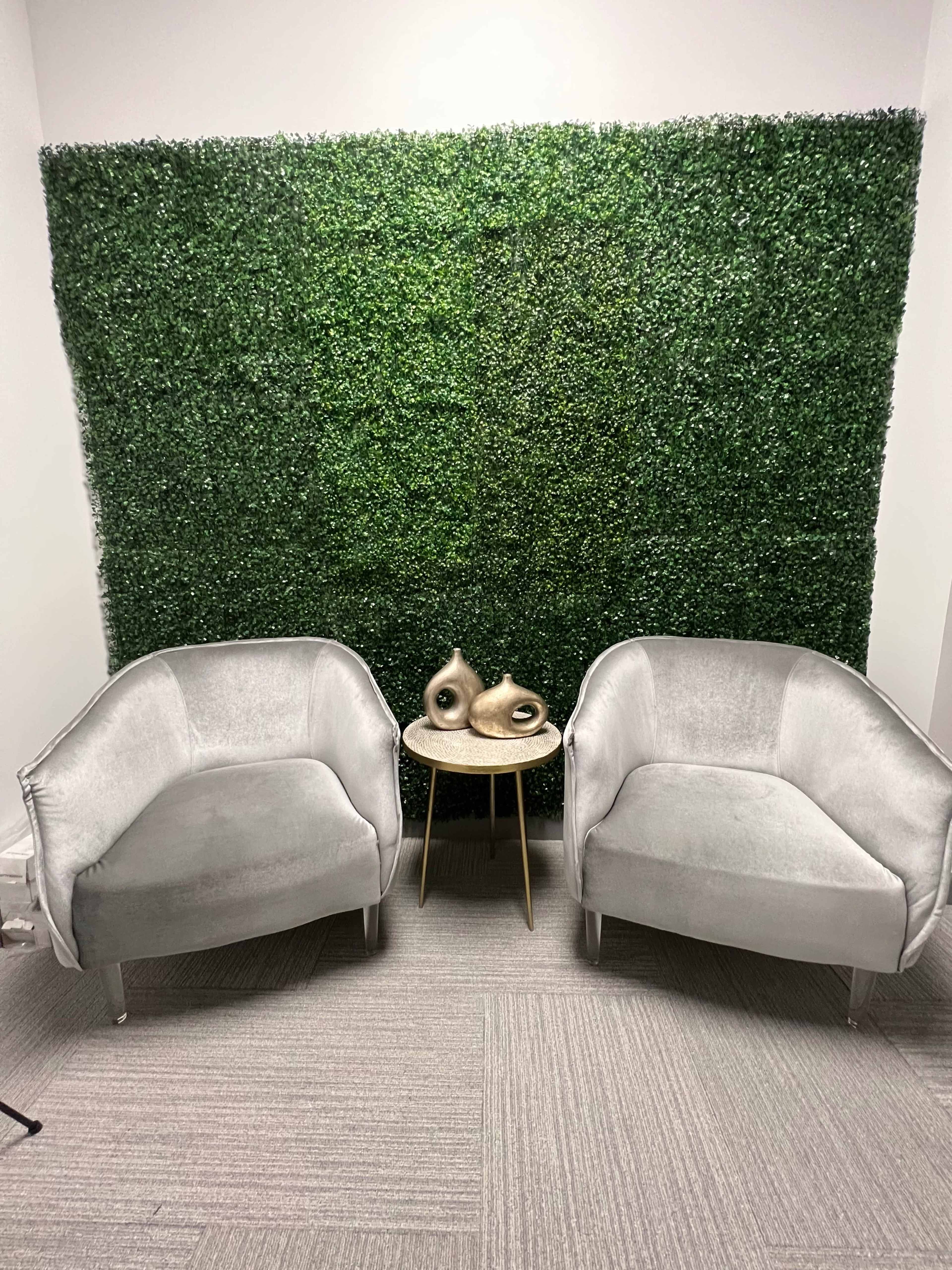 Two gray velvet chairs are positioned opposite each other in front of a green hedge-style wall, with a small round table between them.