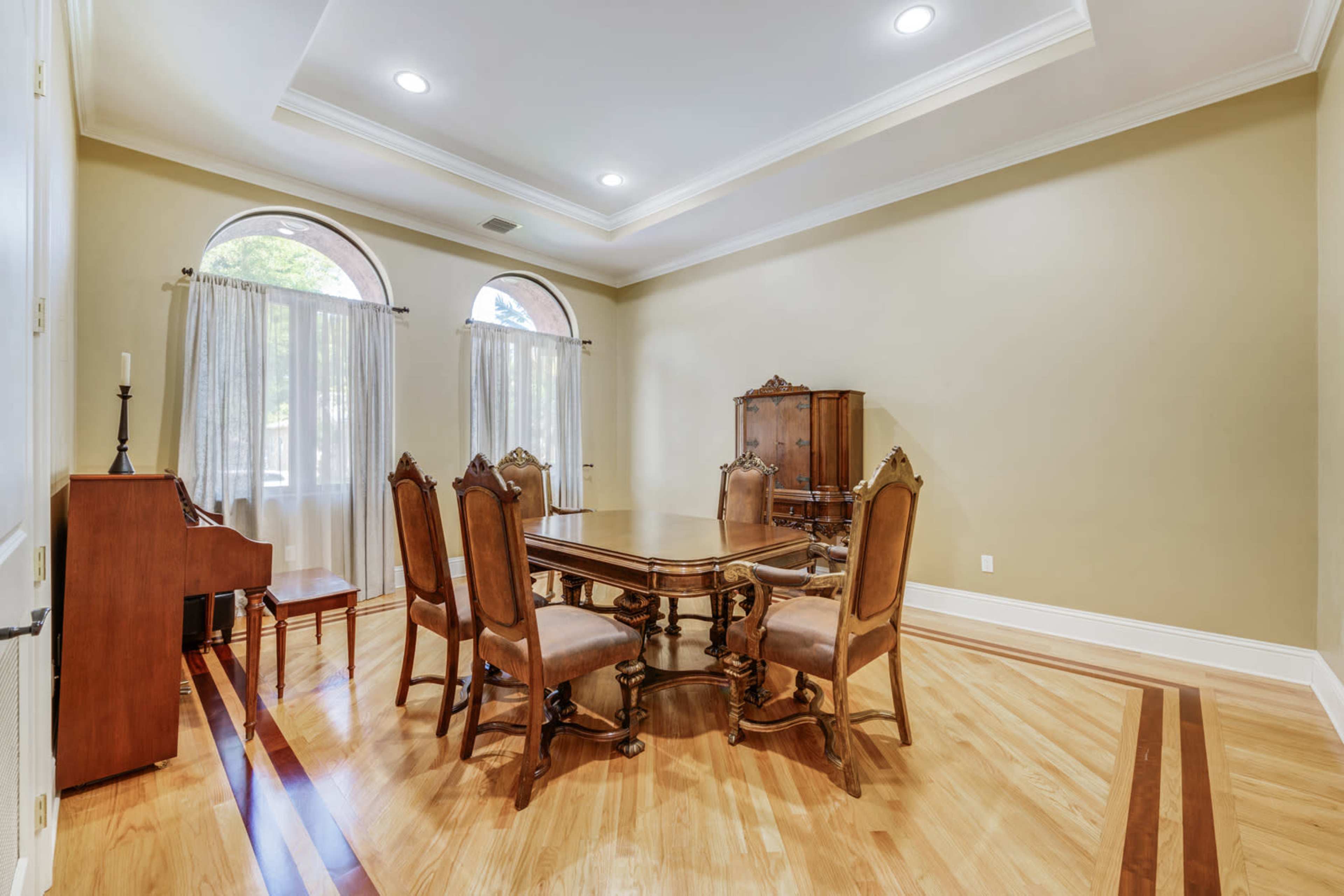 A dining room features a wooden table surrounded by chairs, with large windows draped in sheer curtains and a piano in the corner.