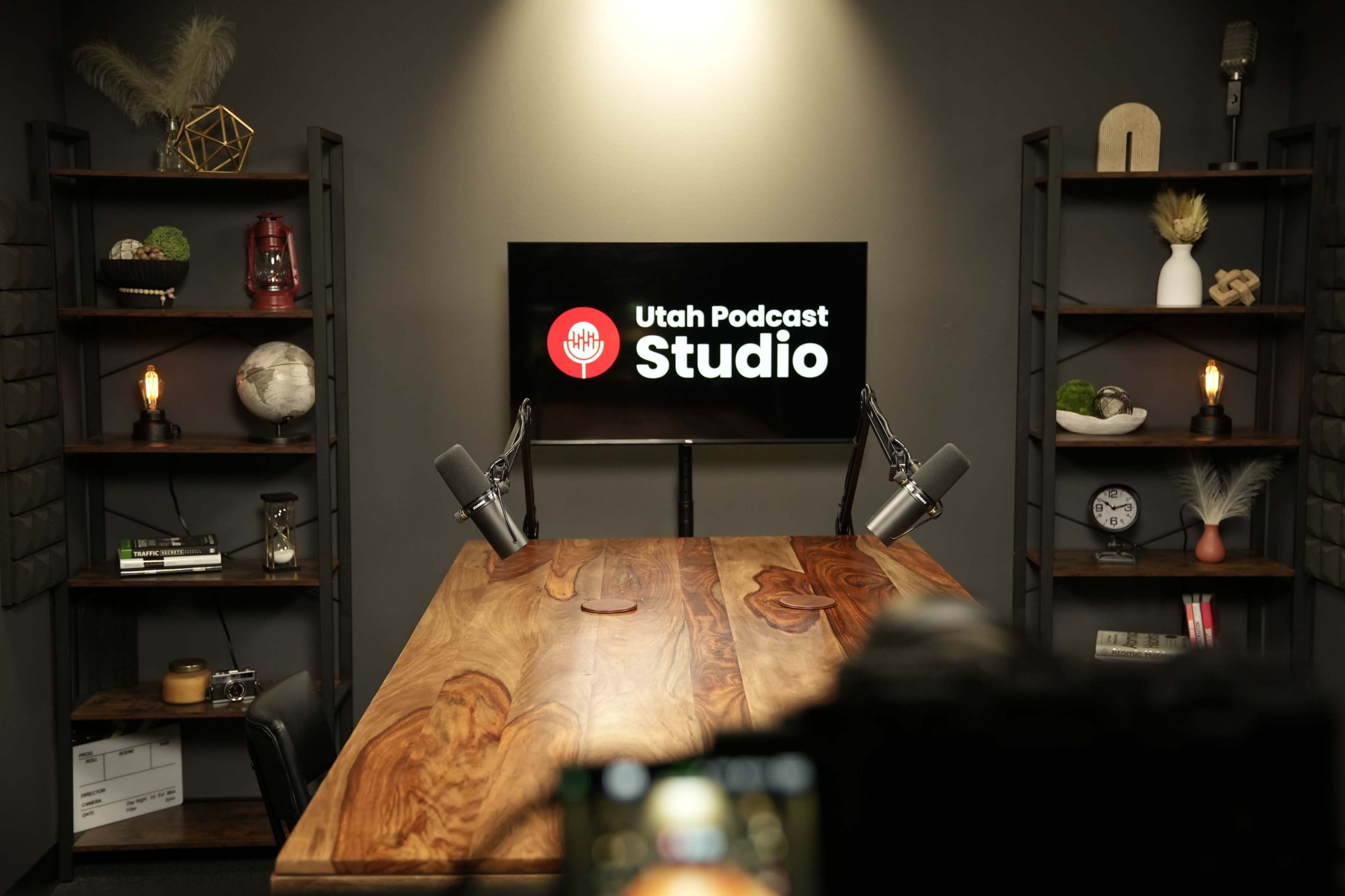 The image shows a podcast studio featuring a wooden table, two microphones, and shelves with various decorative items, all set against a wall displaying the "Utah Podcast Studio" logo on a screen.