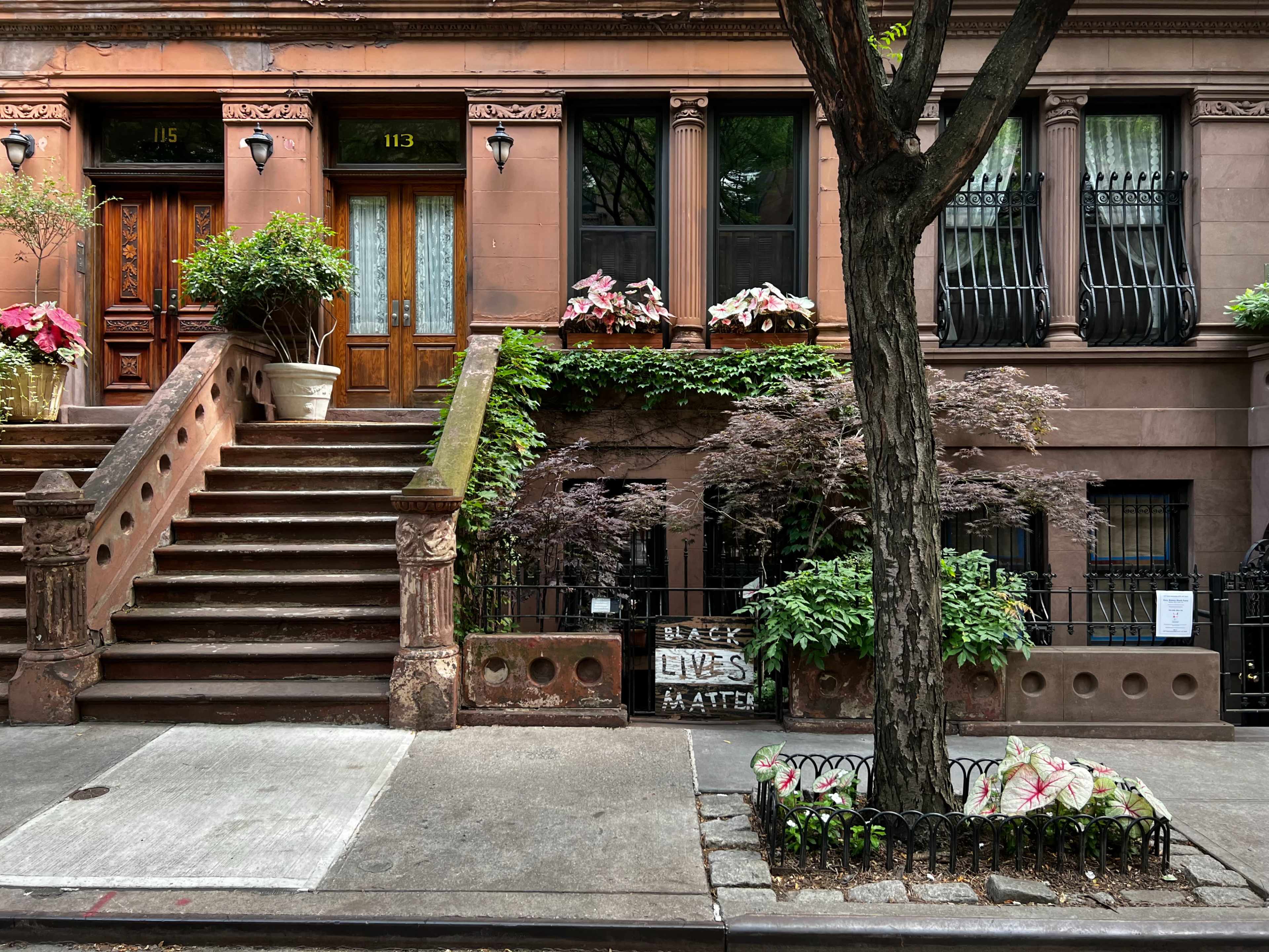 The image shows a brownstone building with a staircase, potted plants, and a "Black Lives Matter" sign in the front yard.