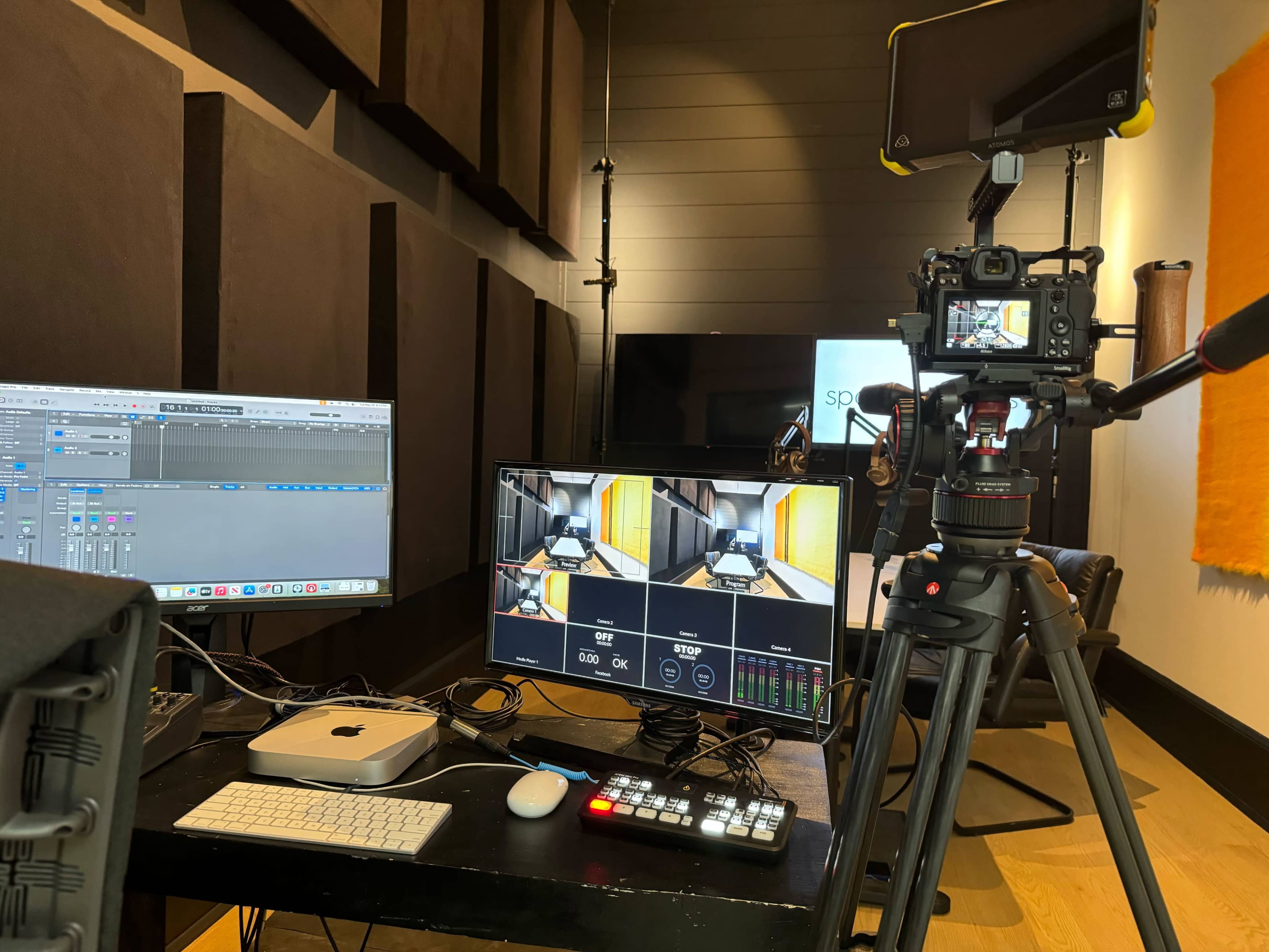 A recording studio setup with two computer monitors, a camera on a tripod, and audio equipment surrounded by soundproofing panels.
