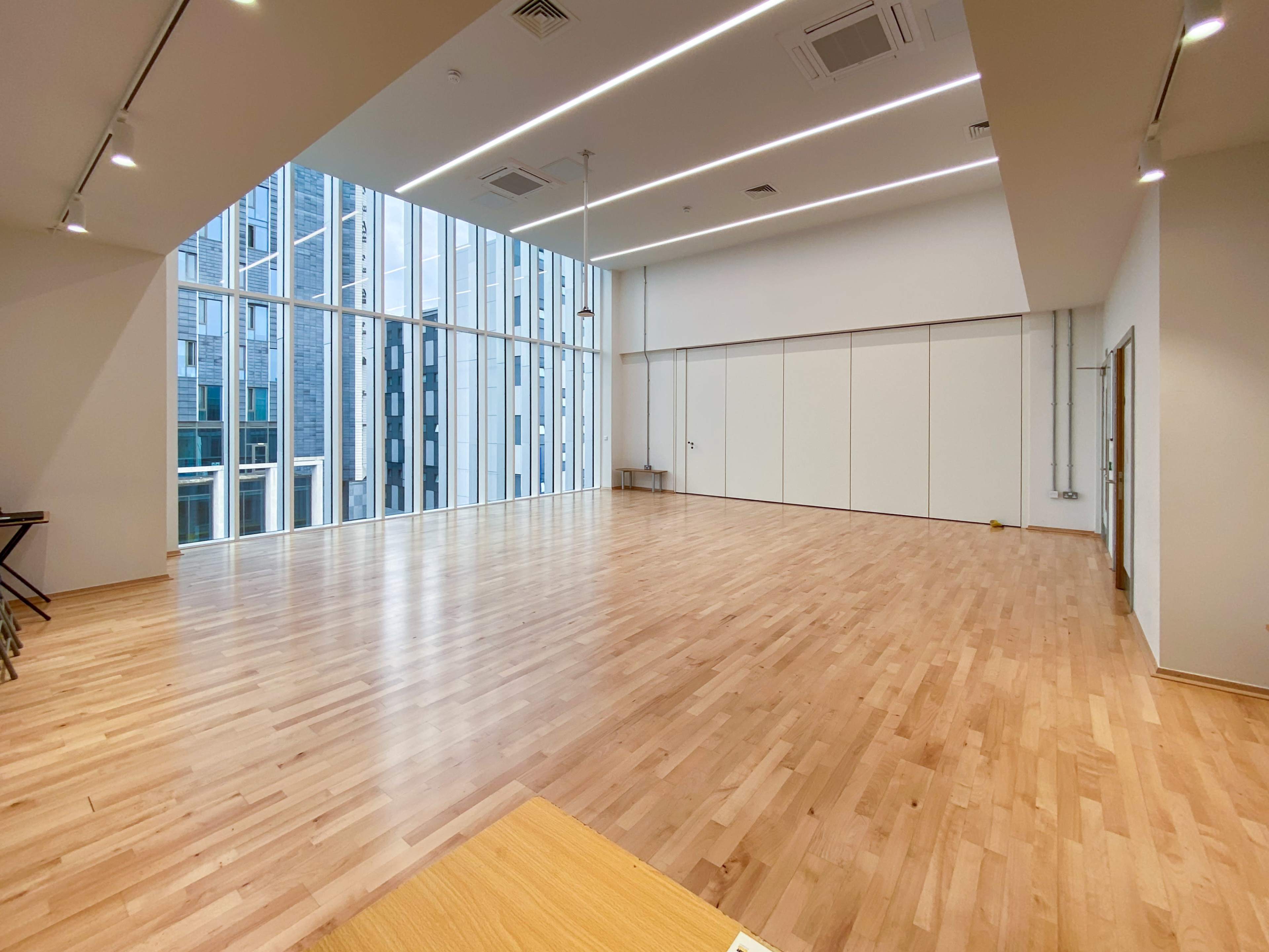 The image shows a spacious, well-lit room with large windows and wooden flooring, featuring minimal furniture.
