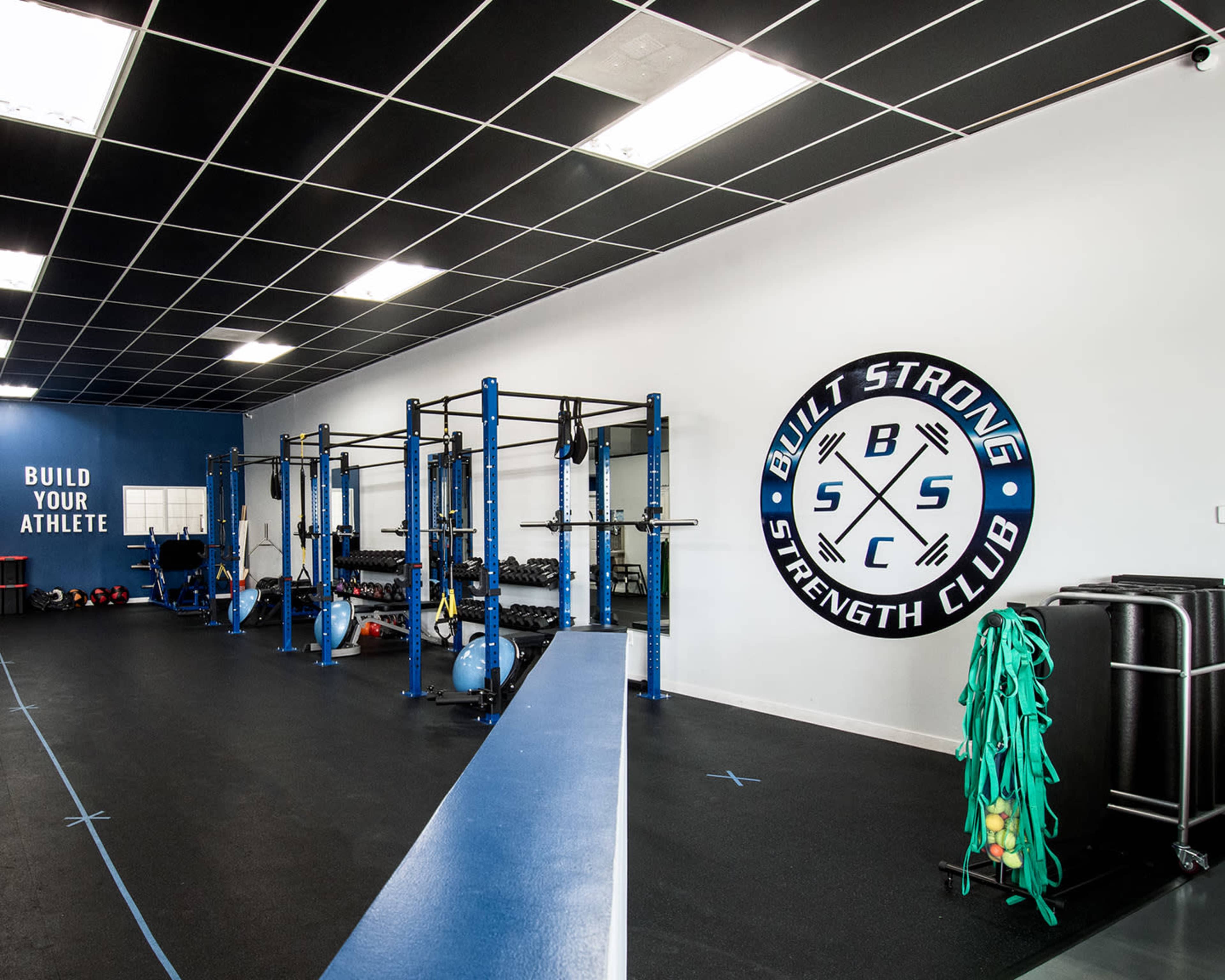 A spacious gym with strength training equipment, including a rack, weights, and exercise balls, against a backdrop of blue and white walls with a logo.