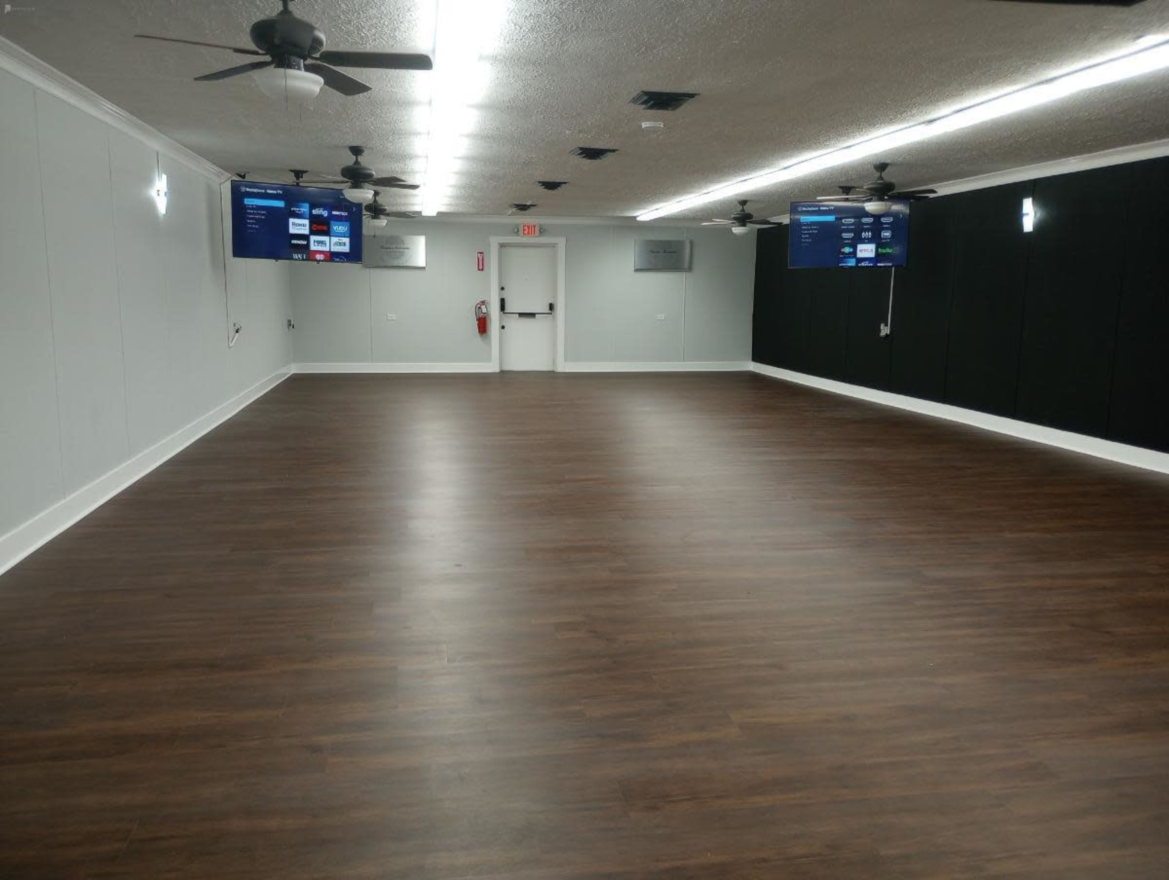 A large, empty room with wooden flooring, two flat-screen televisions mounted on the walls, and ceiling fans.