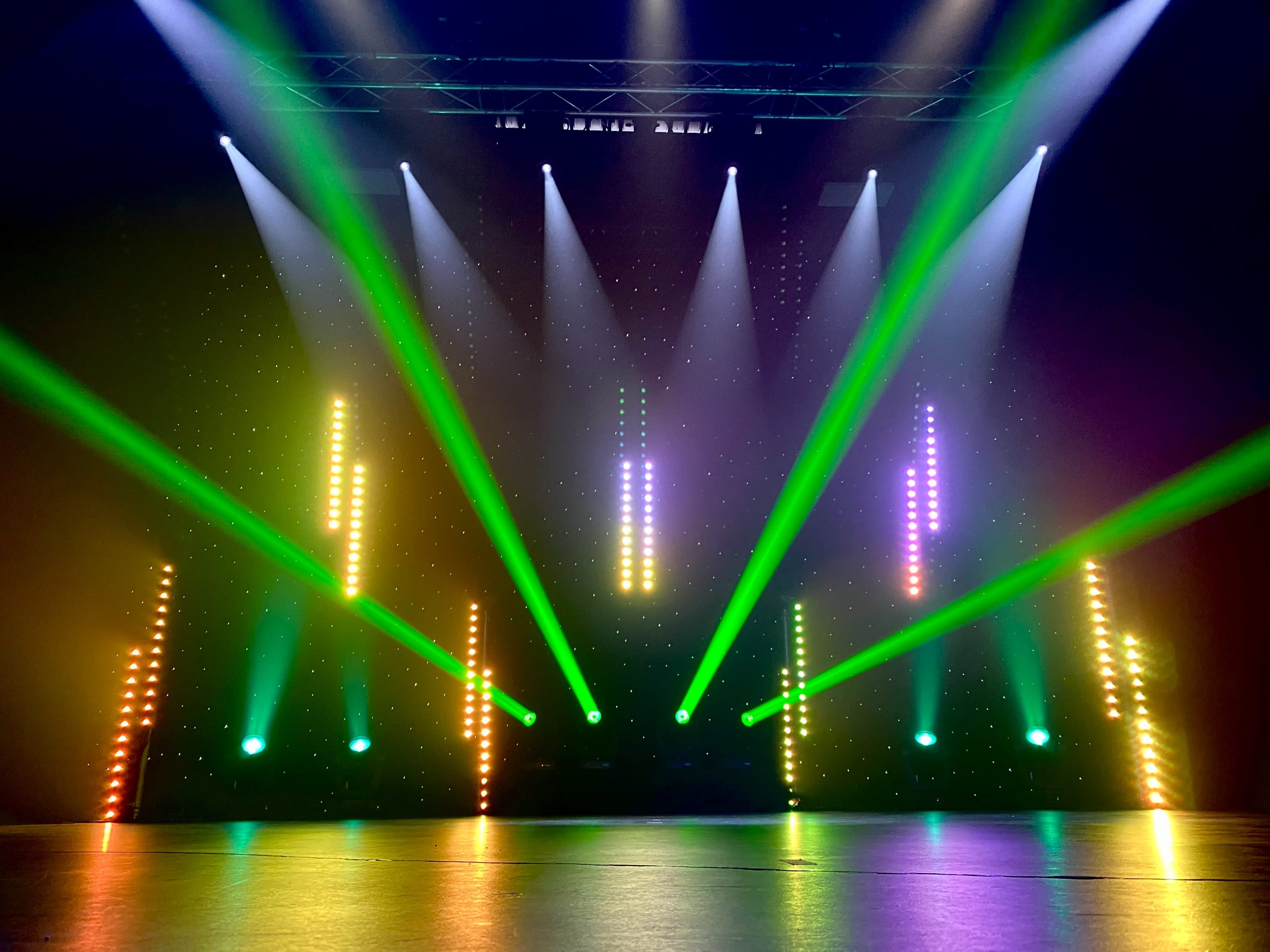 A stage illuminated by vertical light beams in green, yellow, and purple, with a dark backdrop and a shiny floor.