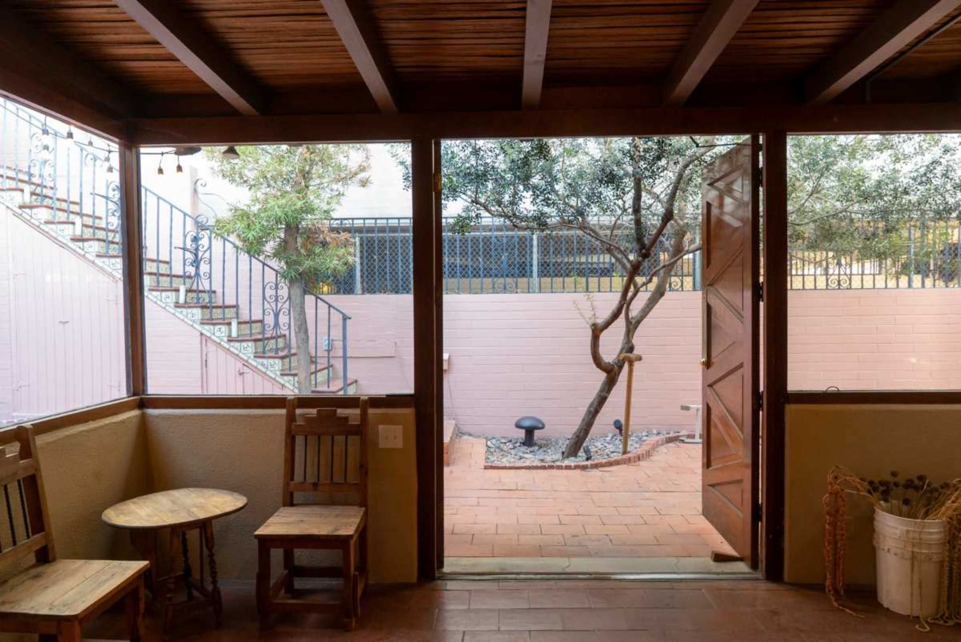 The image shows a wooden interior space with two chairs and a table, leading to a patio with a tree and stairs in the background.