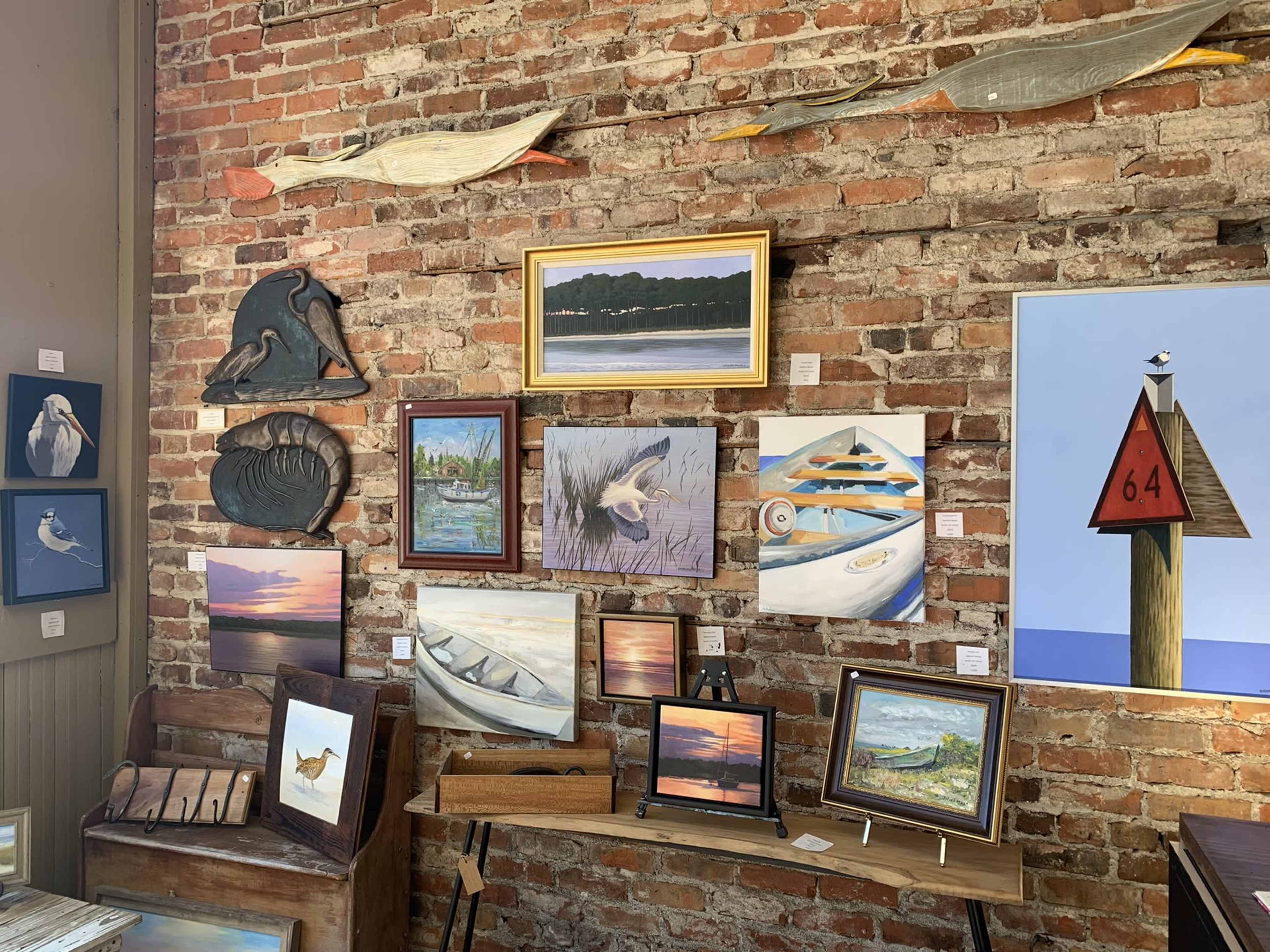 The image shows a brick wall adorned with various paintings and wooden art pieces, alongside a wooden table displaying more artwork.