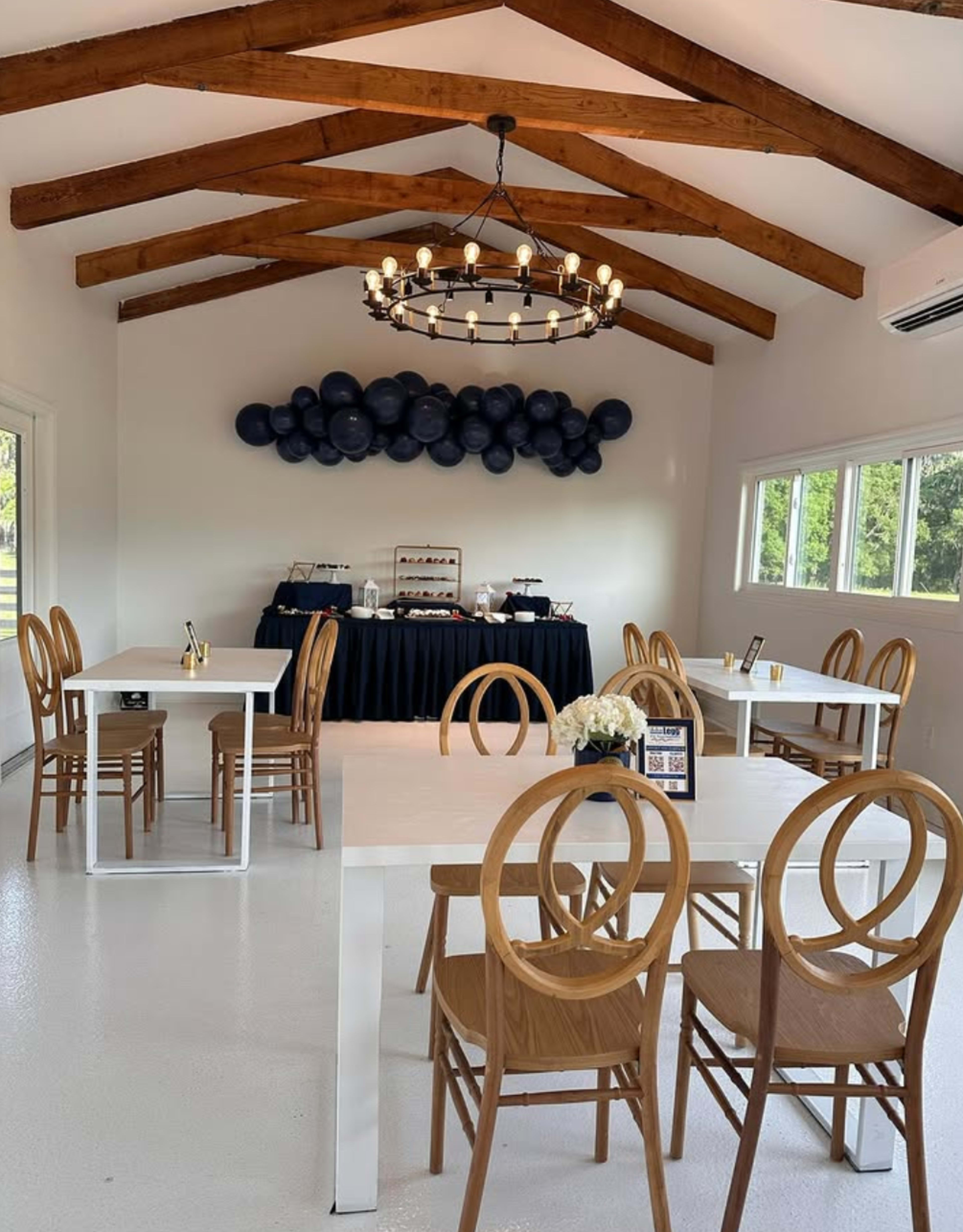 A bright, spacious room features wooden beams, circular wooden tables and chairs, and a decor of blue balloons on the wall against a black table setup.