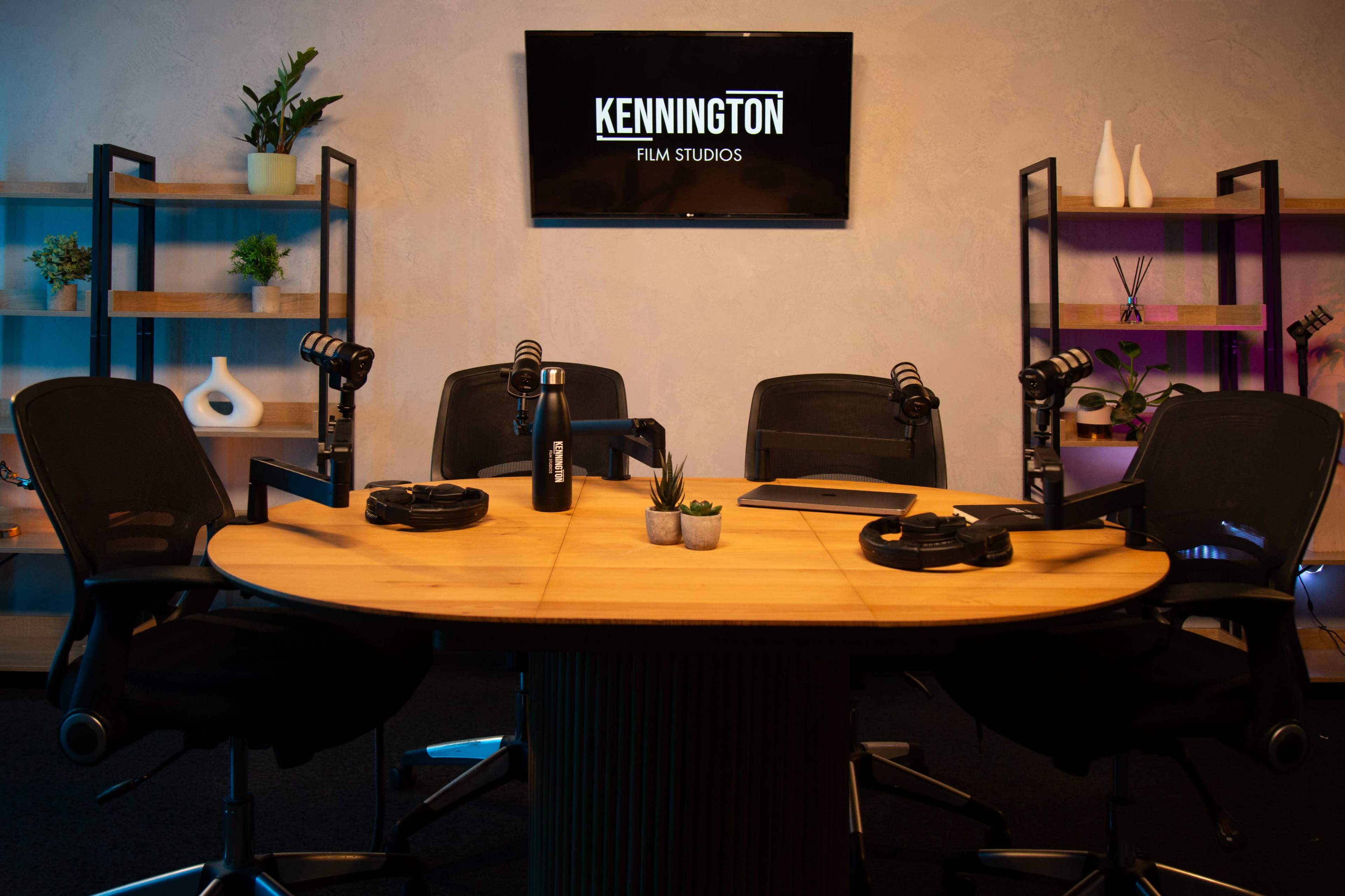 A circular table with microphones and headphones is set up in a studio, featuring a backdrop with the "Kennington Film Studios" logo on a monitor.