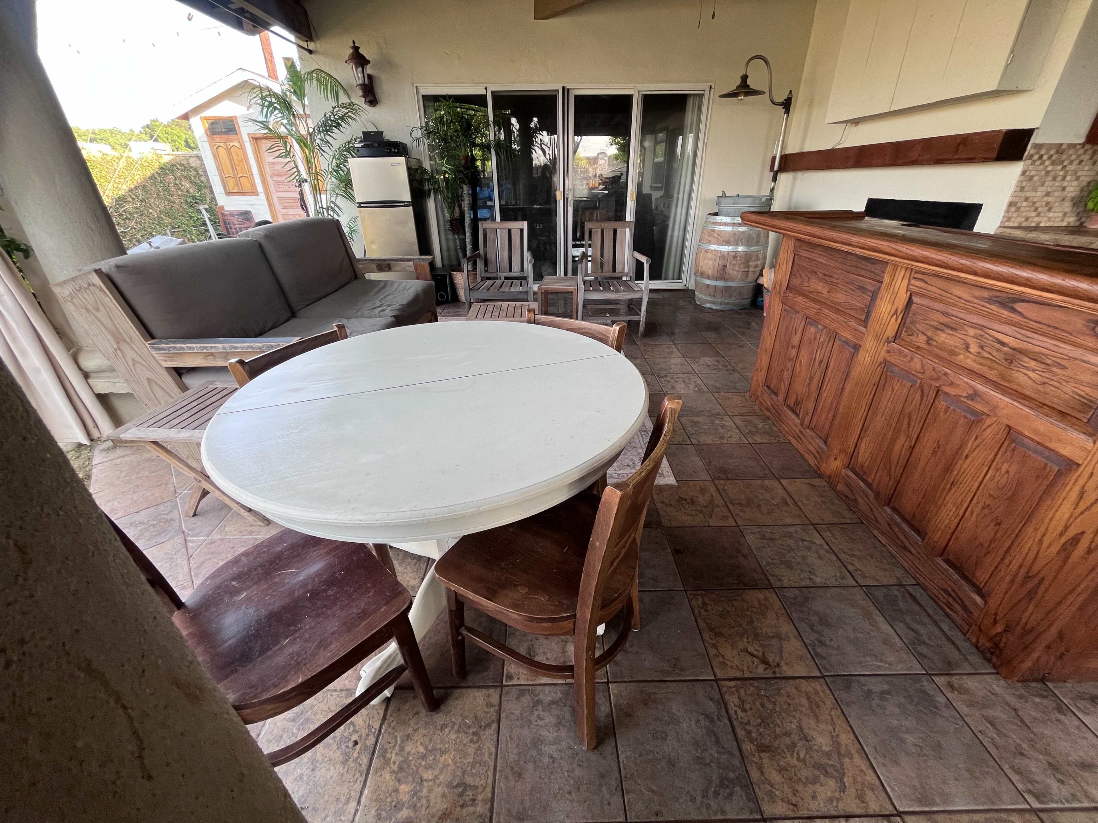 The image shows an outdoor patio area with a round white table surrounded by four wooden chairs, a seating area with a gray couch, and a wooden bar counter.