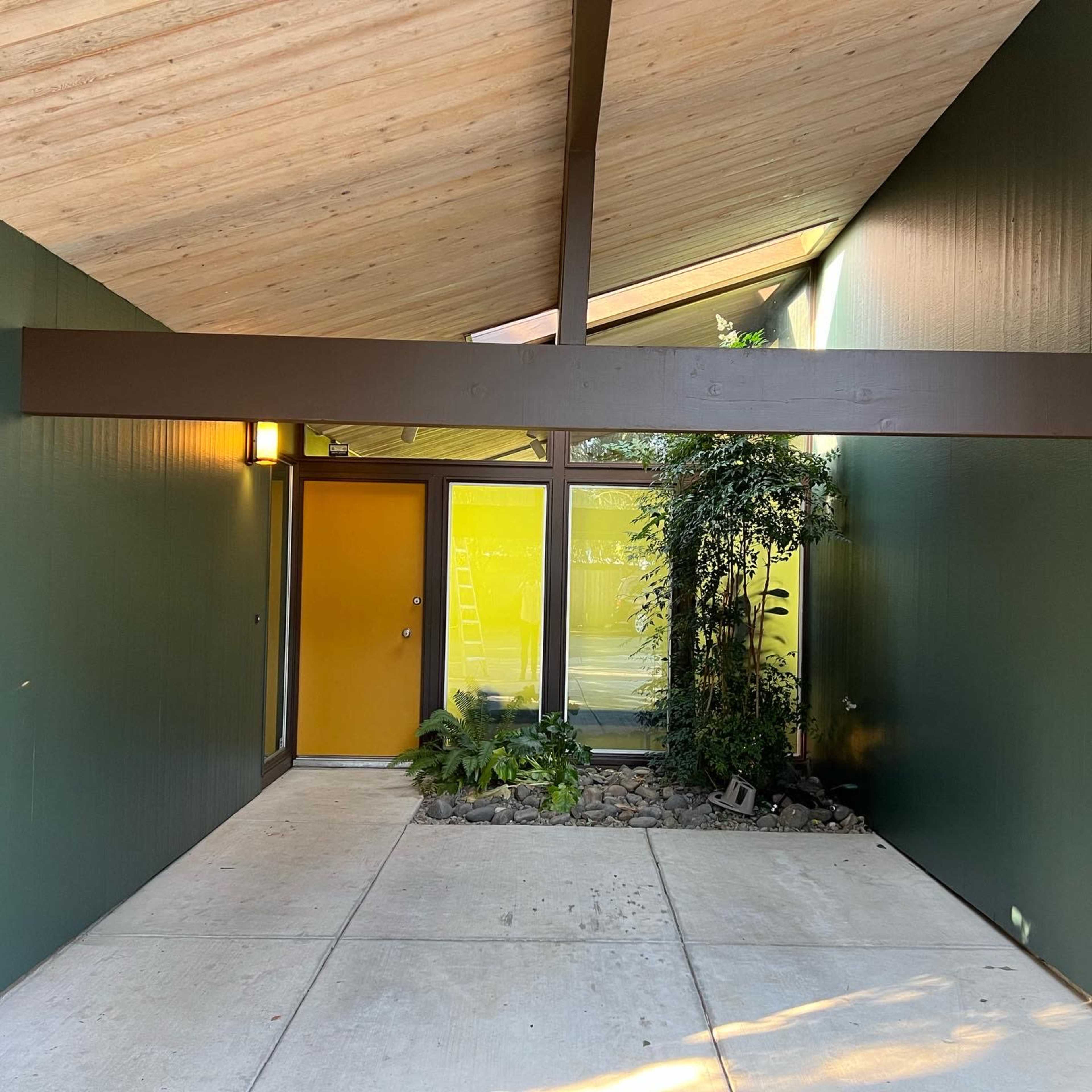 An exterior entrance of a modern building with a sloped roof and a yellow door, framed by dark green walls and a landscaped area featuring rocks and plants.
