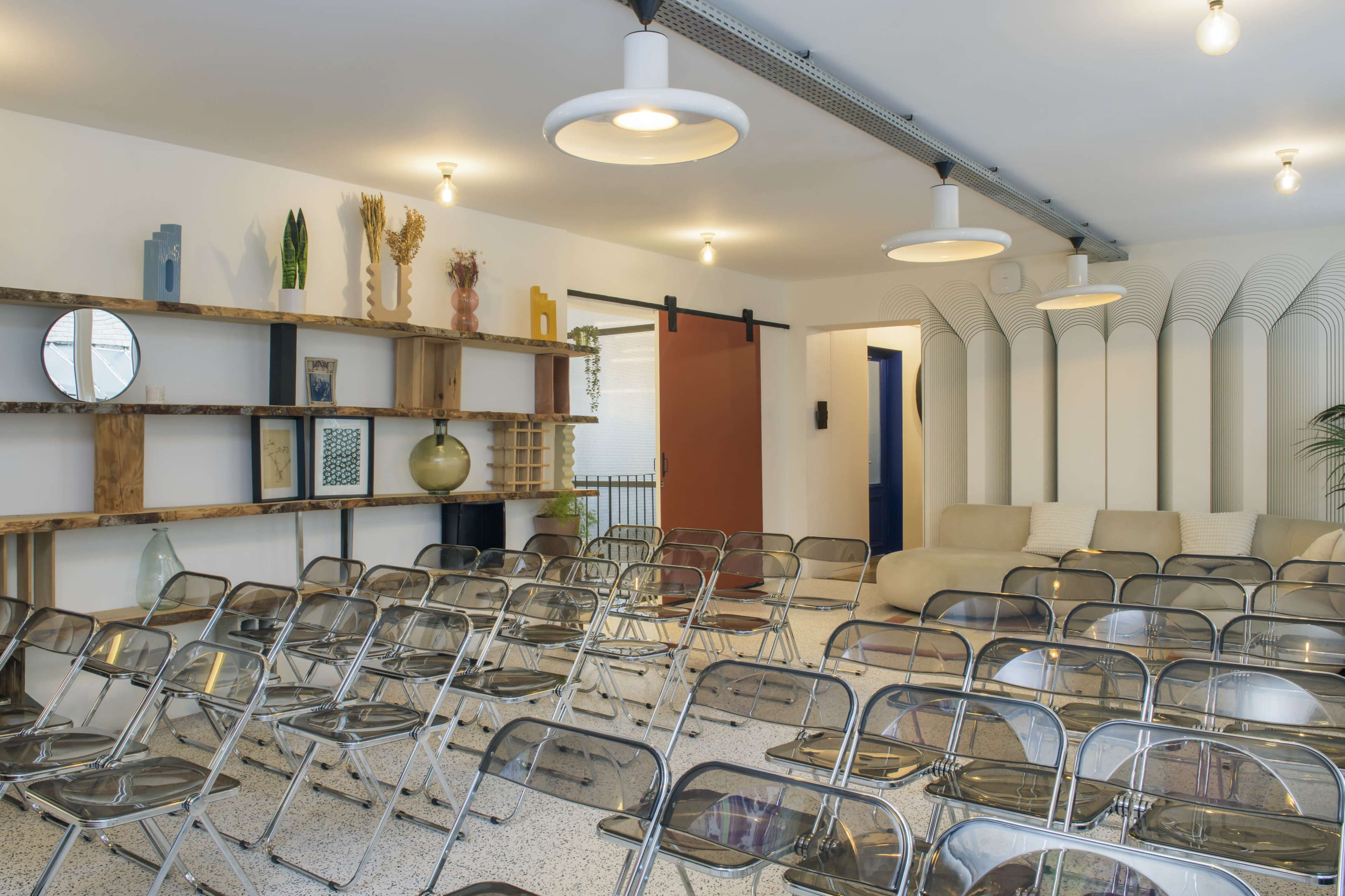 The image depicts a modern meeting room with rows of clear plastic chairs facing a wall-mounted shelf featuring decorative items, alongside a cozy seating area.