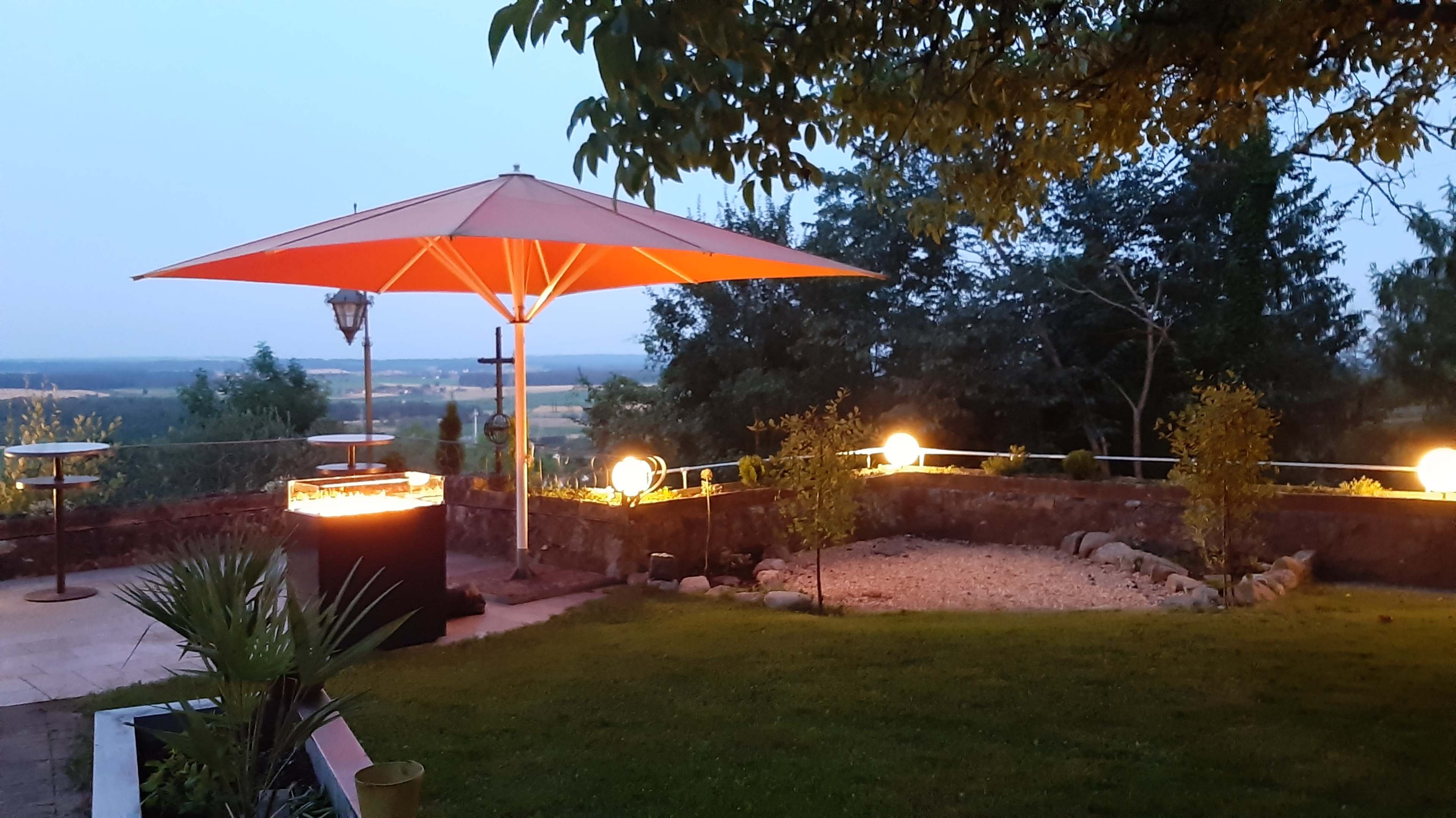 A patio scene features an orange umbrella over a grill, surrounded by landscaped greenery and glowing orbs of light in the dusk.