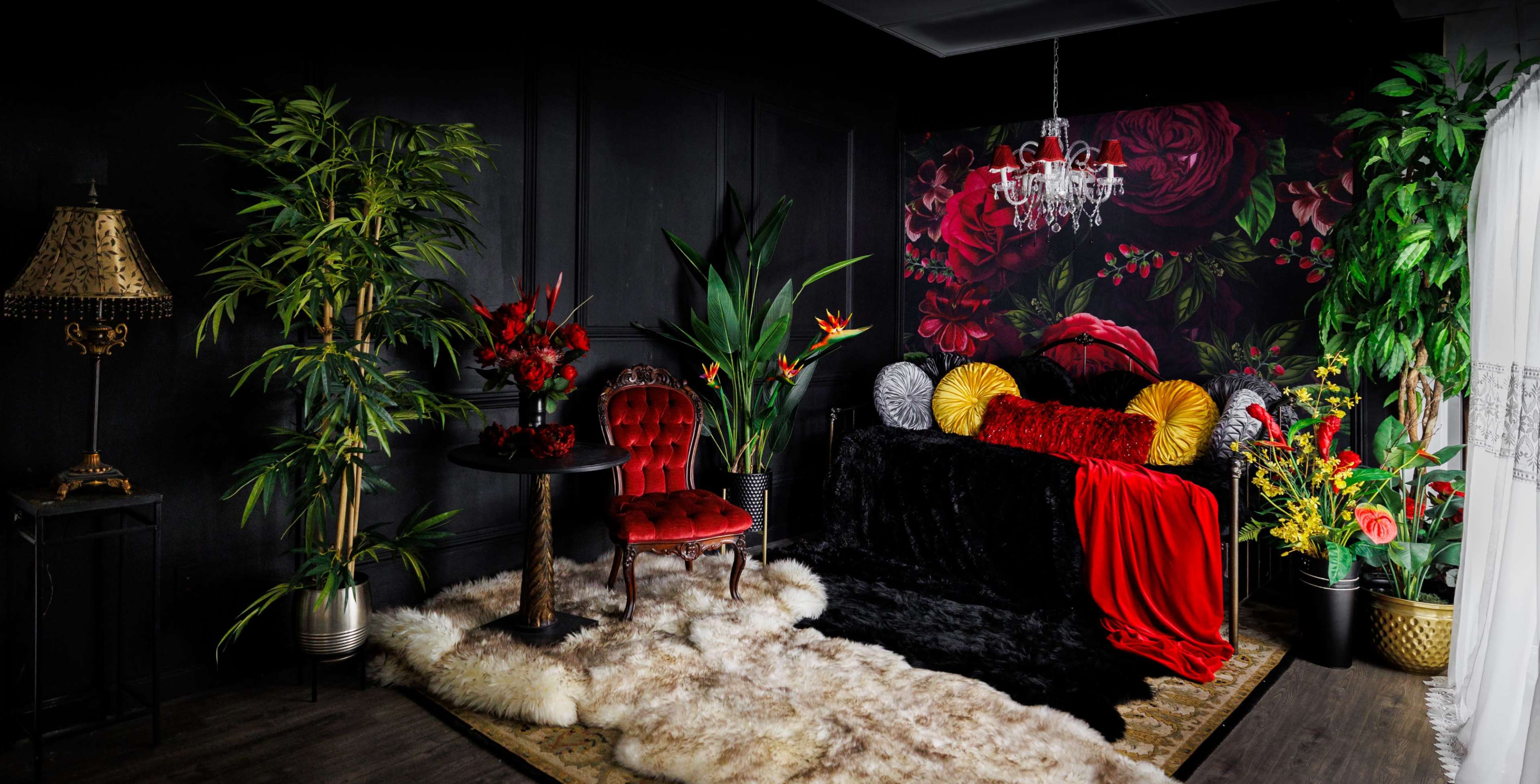 The image shows a dark, elegantly styled room featuring a plush black couch adorned with colorful pillows, a vintage red chair, a decorative table, and various tropical plants against a floral backdrop.