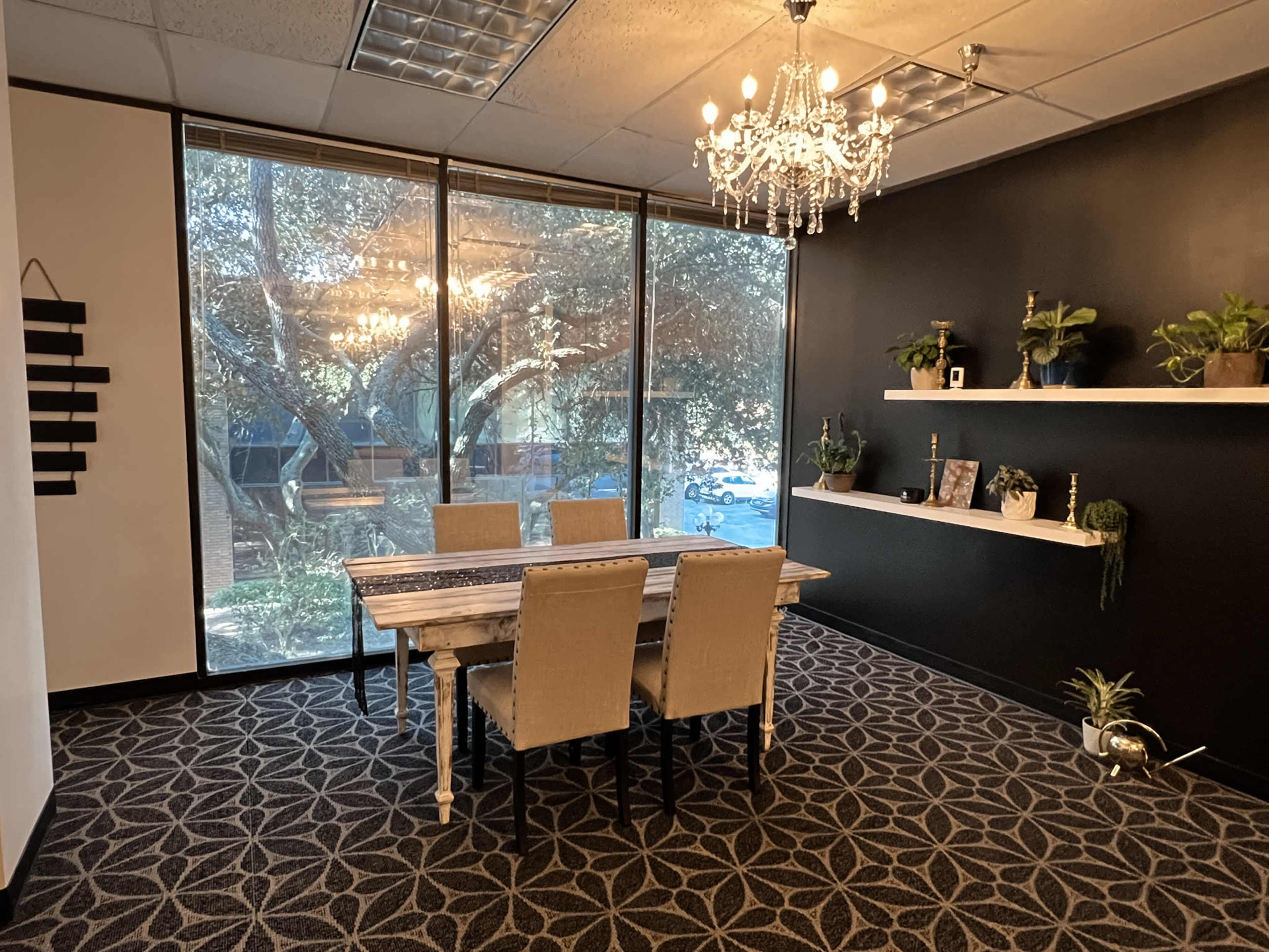 A modern meeting room with a wooden table surrounded by four beige chairs, large windows providing natural light, and a chandelier hanging from the ceiling.