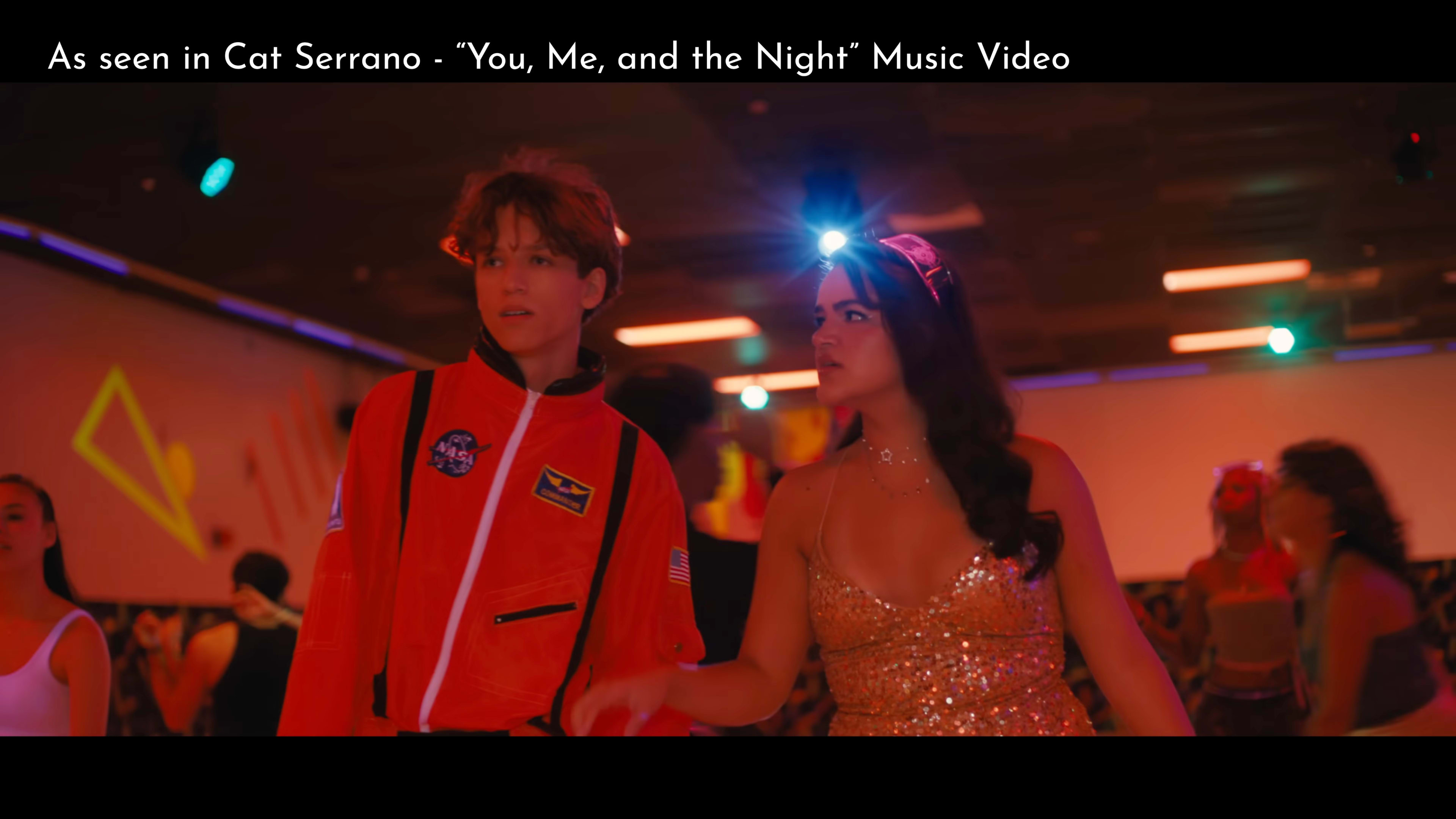 The image shows two young people in a vibrant roller rink setting, with one in an orange jacket and the other in a glittering dress, as they appear to be interacting amidst a lively crowd.