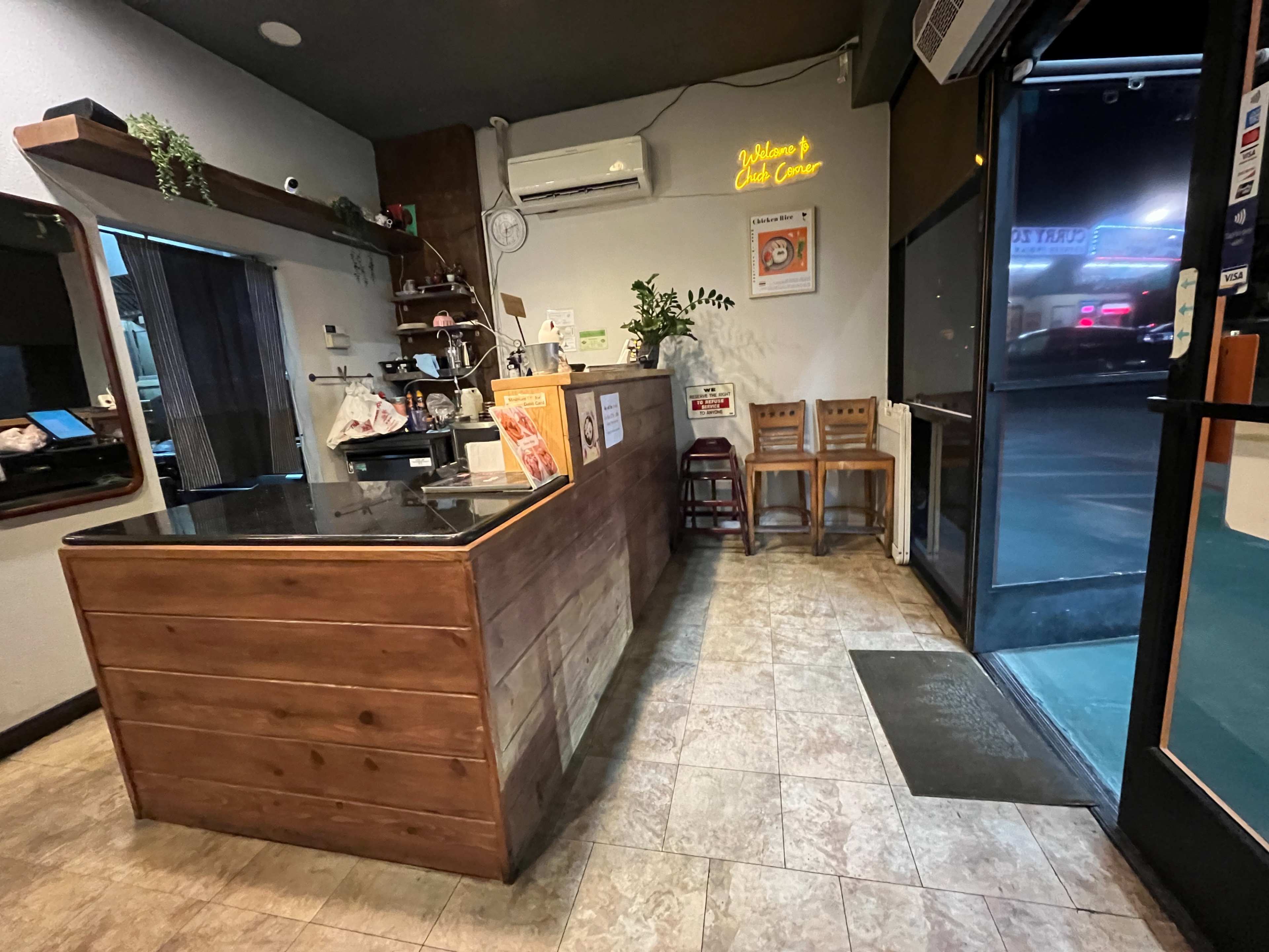 The image shows the interior of a small food establishment with a wooden counter, a few chairs, and a door leading outside.