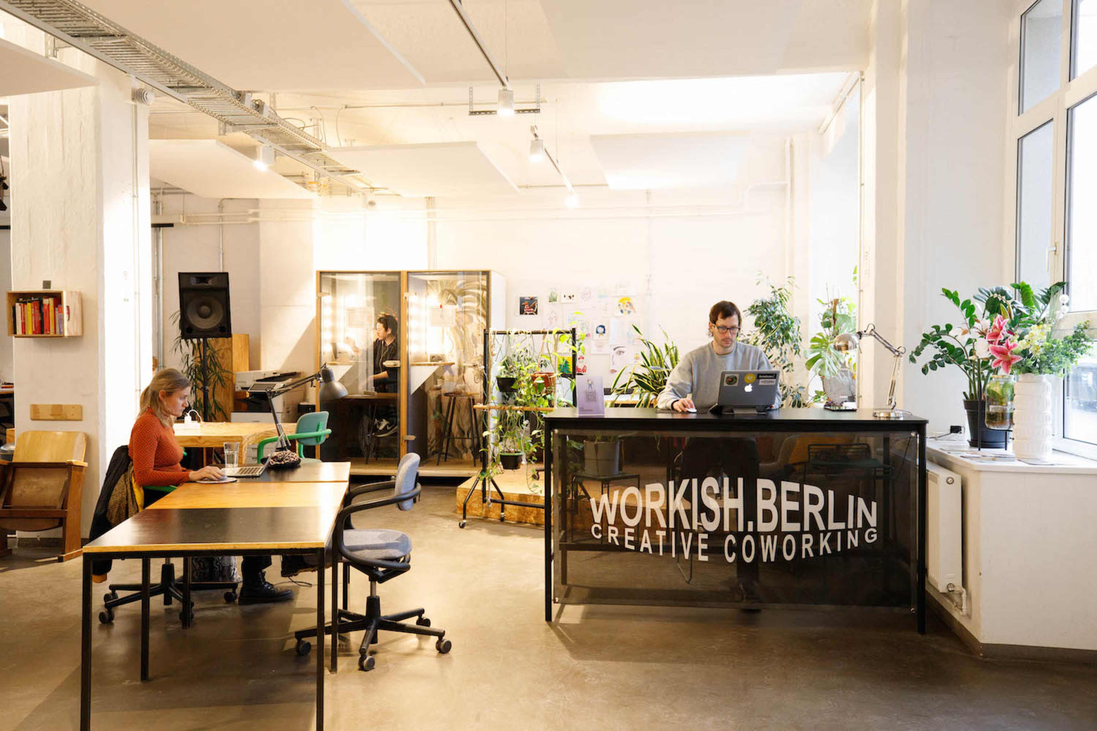 A modern coworking space features a glass reception desk, plants throughout, and individuals working at tables.