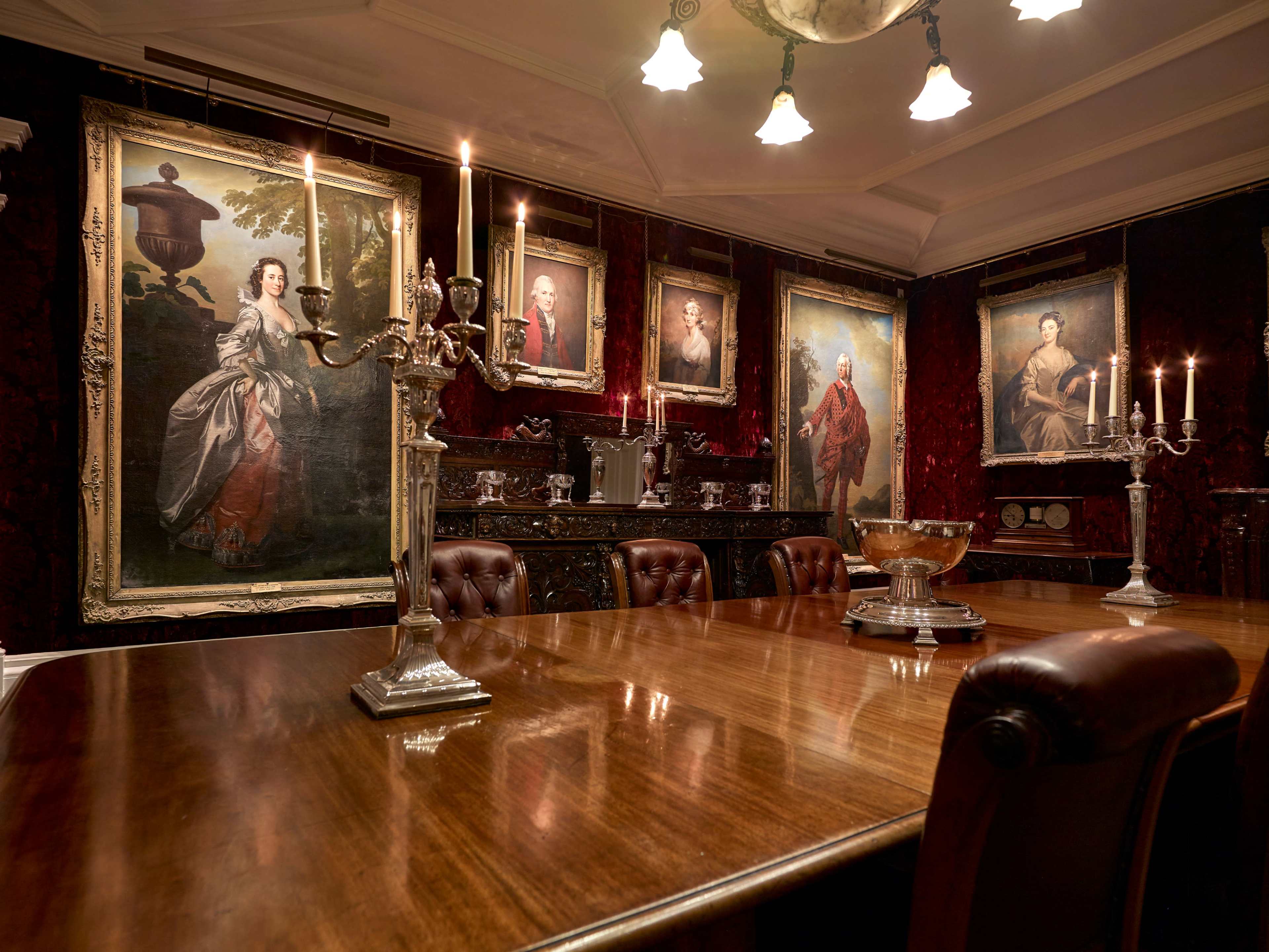 The image shows an elegant dining room with a long wooden table, Victorian-style portraits on the walls, and silver candelabras providing illumination.