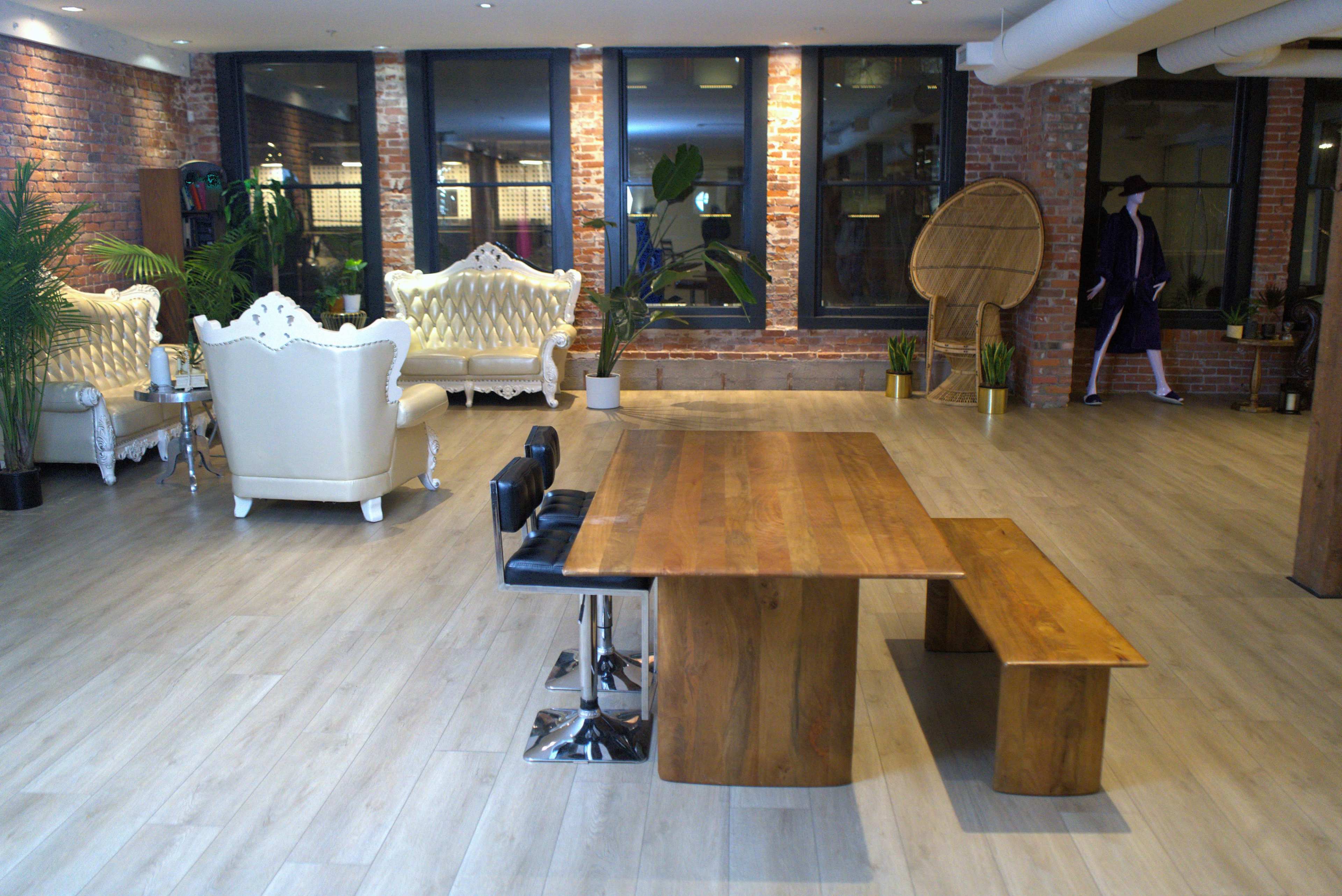 The image shows a spacious room with wooden flooring, featuring a large wooden table and bench, elegant white couches, and greenery, along with exposed brick walls and large windows.