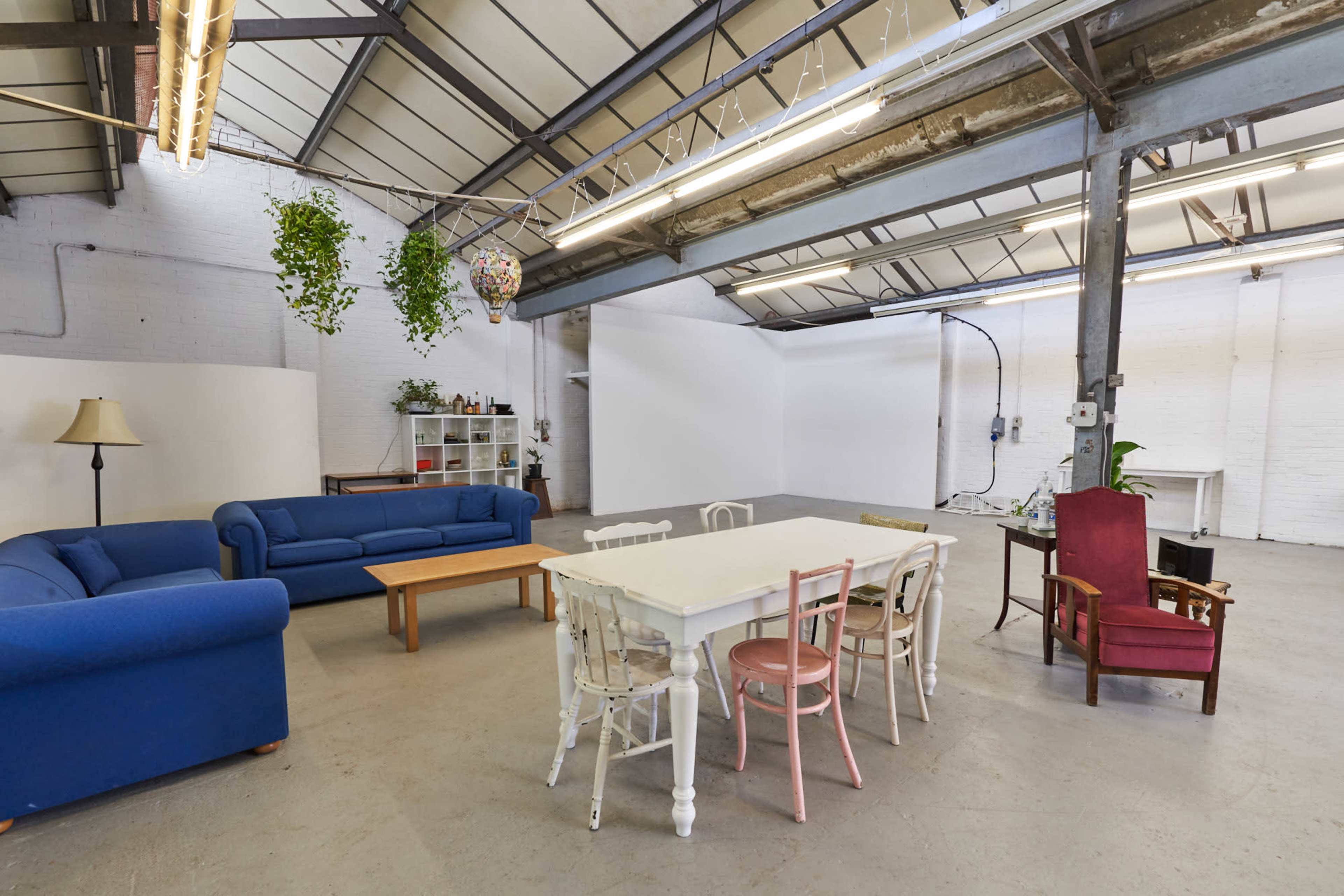 A spacious, industrial-style room with a blue sofa, a wooden coffee table, a white dining table surrounded by chairs, and various plants hanging from the ceiling.