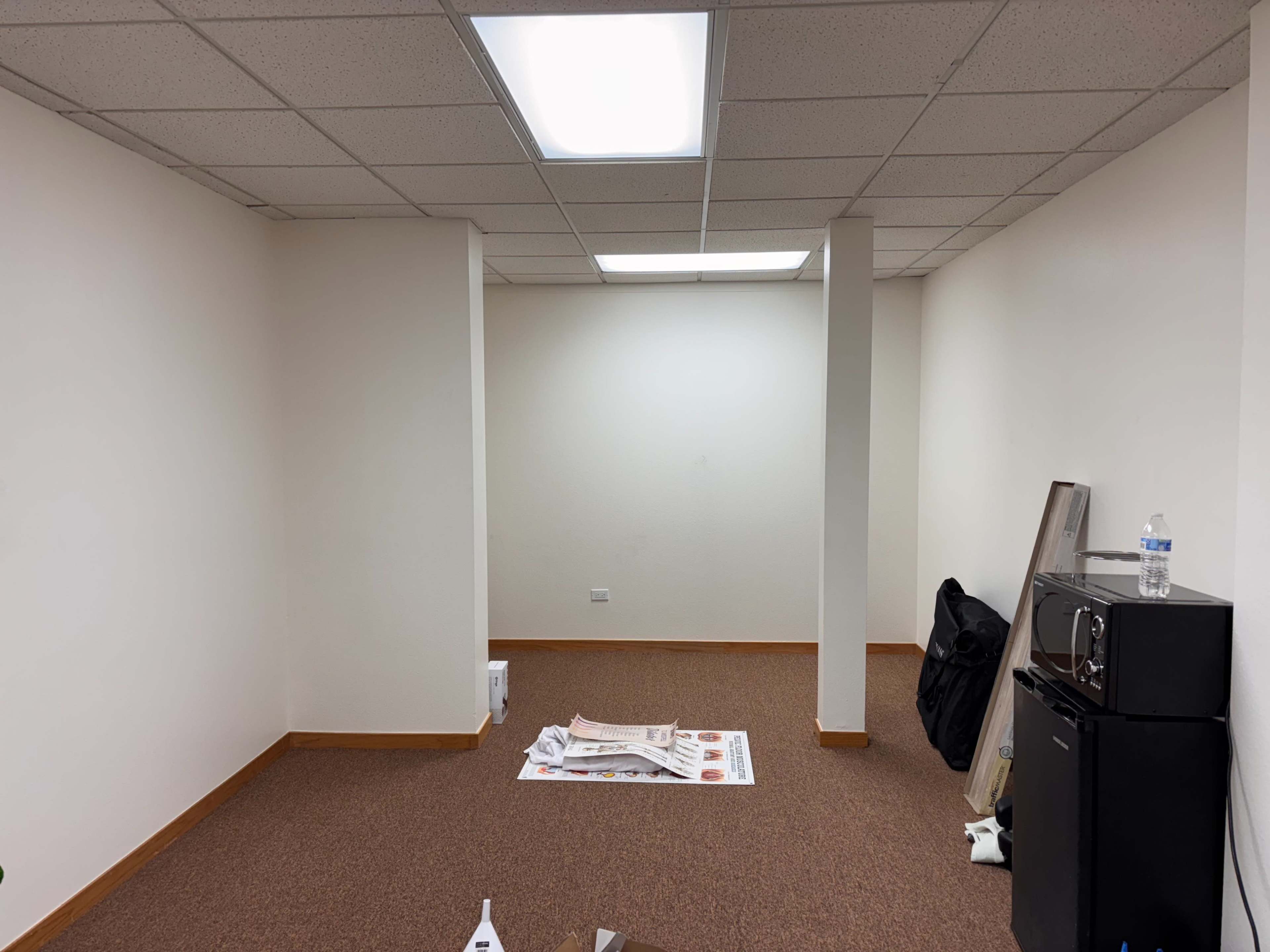 The image shows a sparsely furnished room with beige walls, carpeted flooring, and a few items such as a water bottle and papers on the floor.