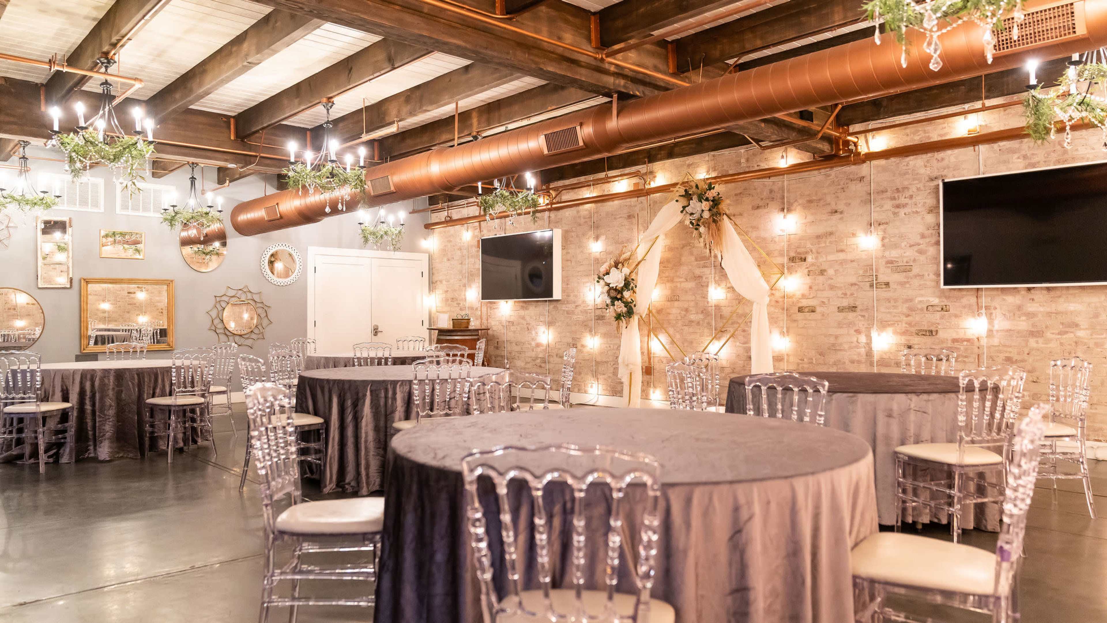 A decorated event space featuring round tables with gray tablecloths, clear acrylic chairs, and walls adorned with mirrors and soft lighting.