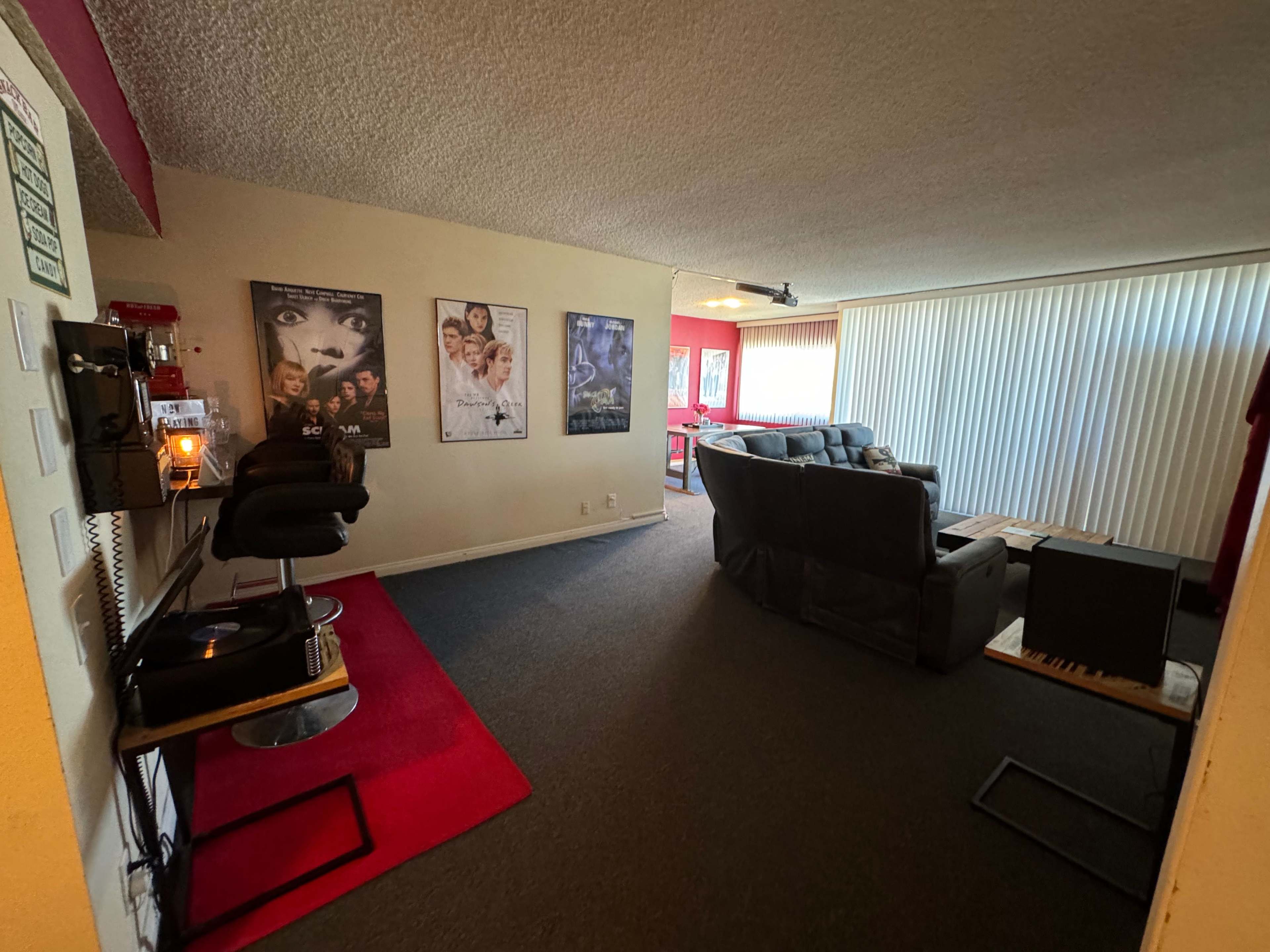 The image shows a living space featuring a seating area with a large black couch, a small waiting area with a chair, and movie posters on one wall.