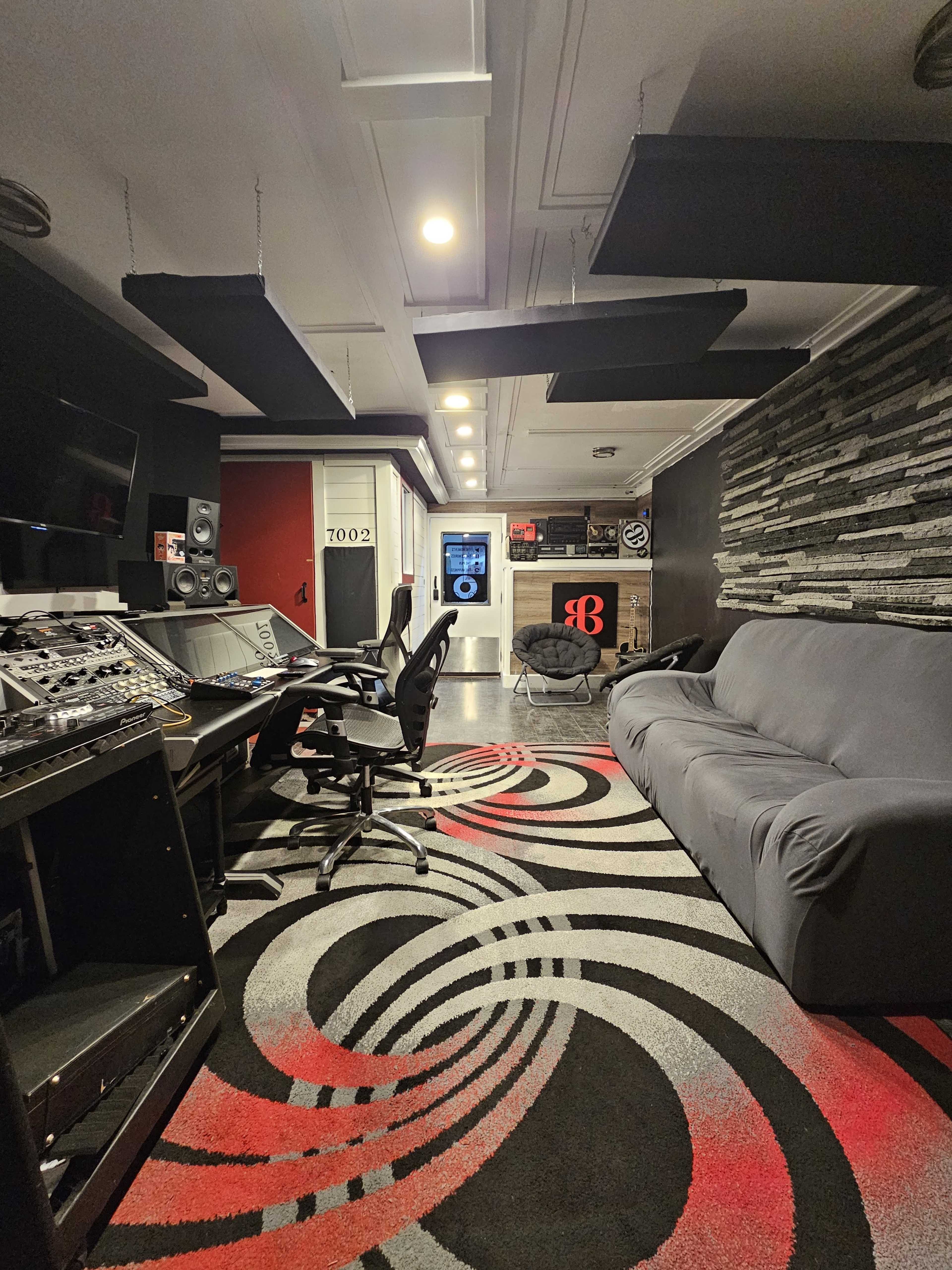 The image shows a modern recording studio with a control desk, black and red accents, and a gray couch, featuring soundproofing panels and a patterned carpet.