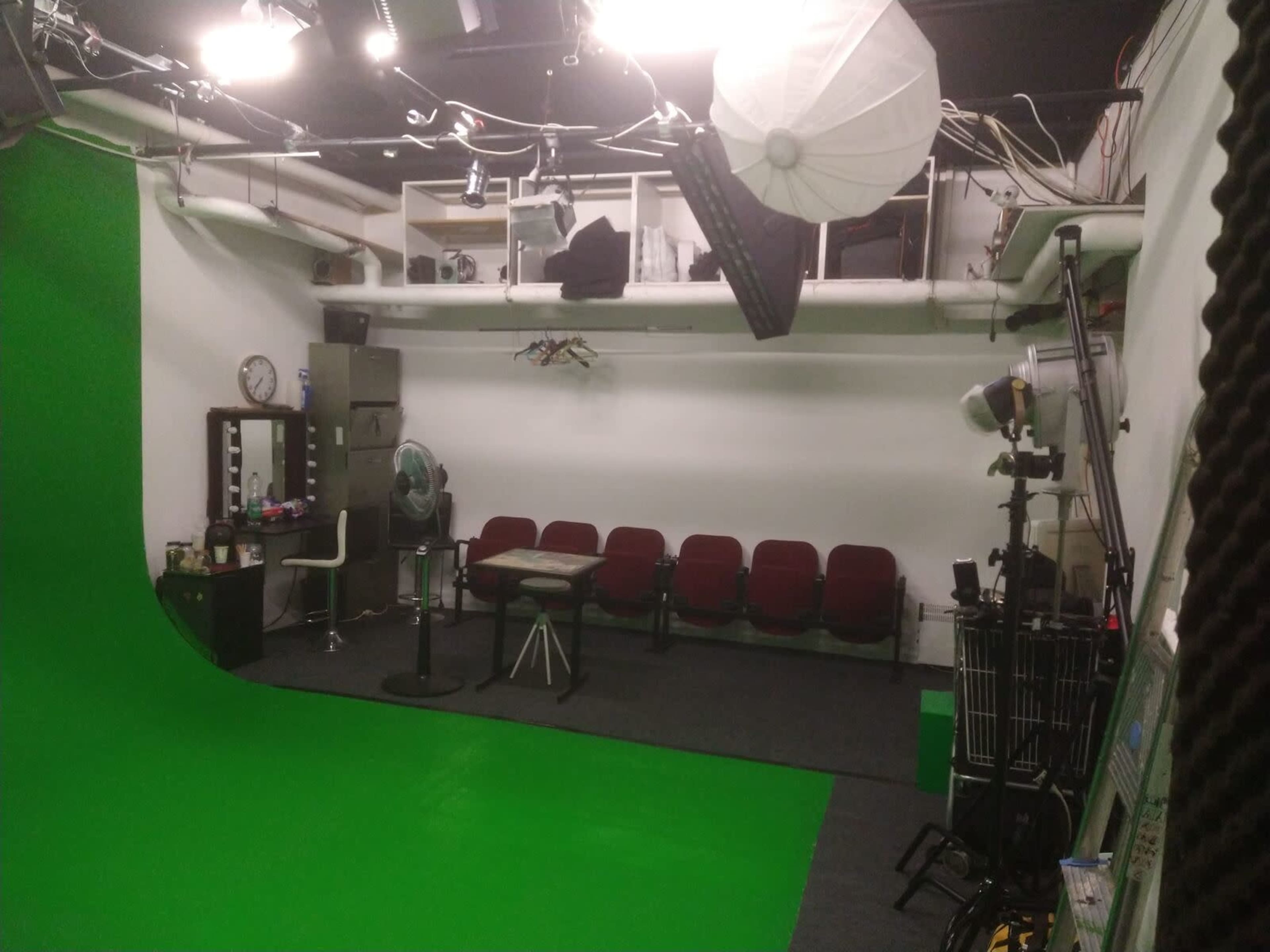 The image depicts a green screen studio with an empty seating area, a table, and various lighting equipment.