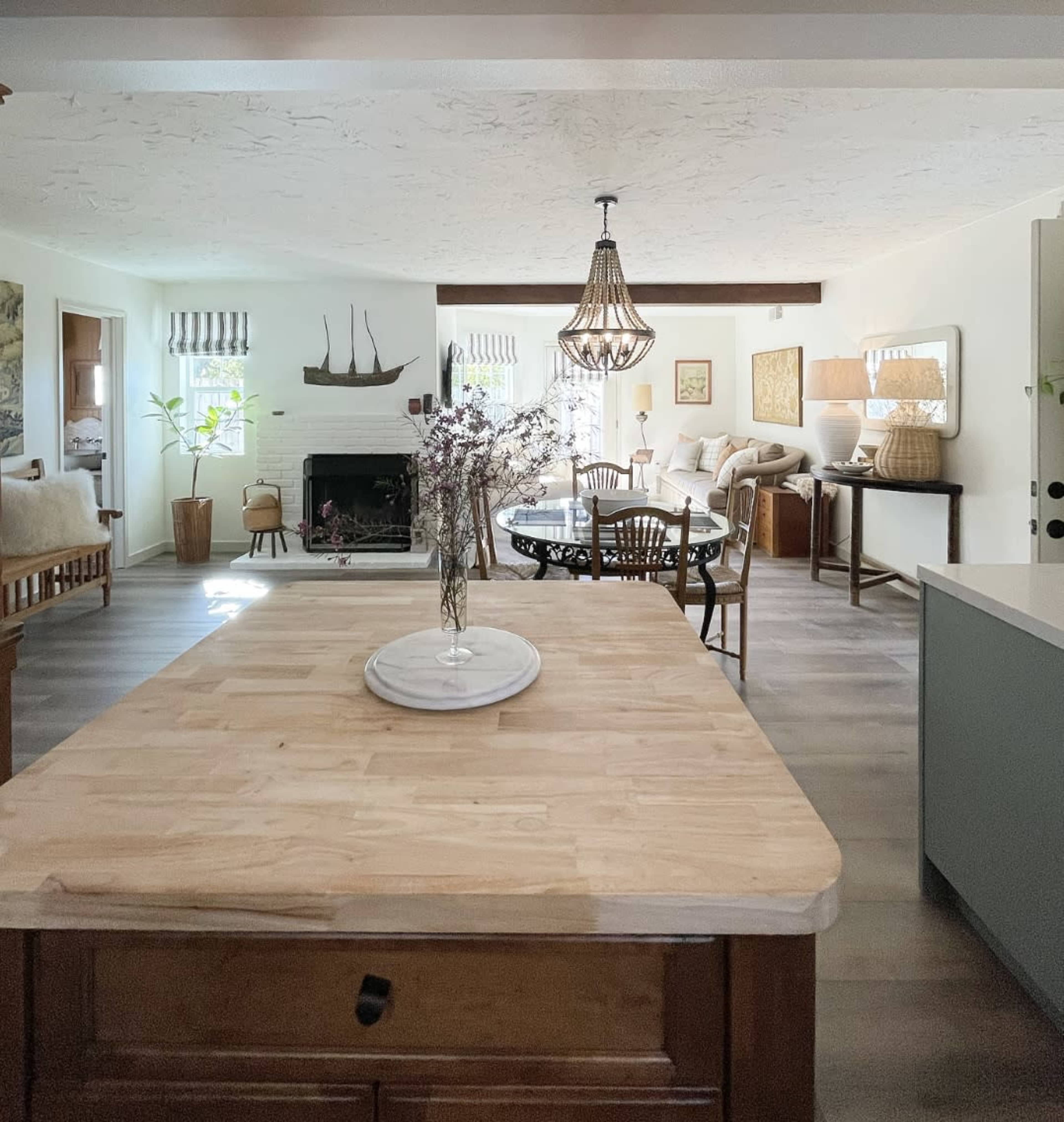 A wooden kitchen island is centered in a brightly lit open-concept living space that includes a dining area, seating area, and a fireplace.