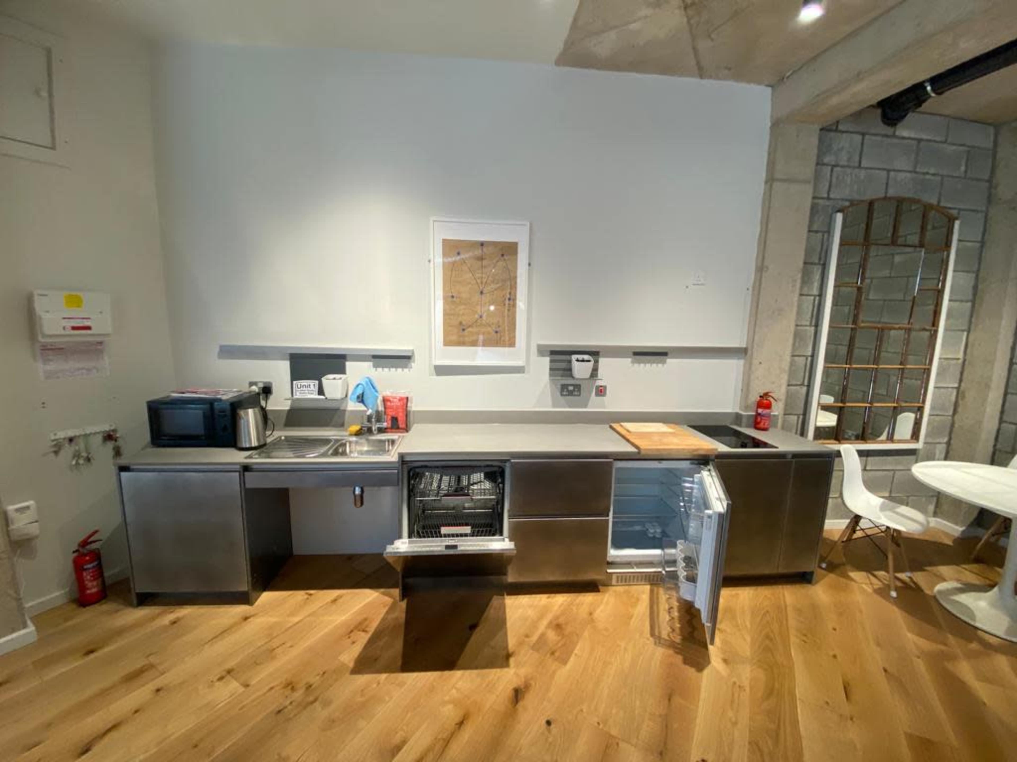 A modern kitchen features stainless steel cabinets, a sink, a microwave, and an open dishwasher, with wooden flooring and a framed map on the wall.