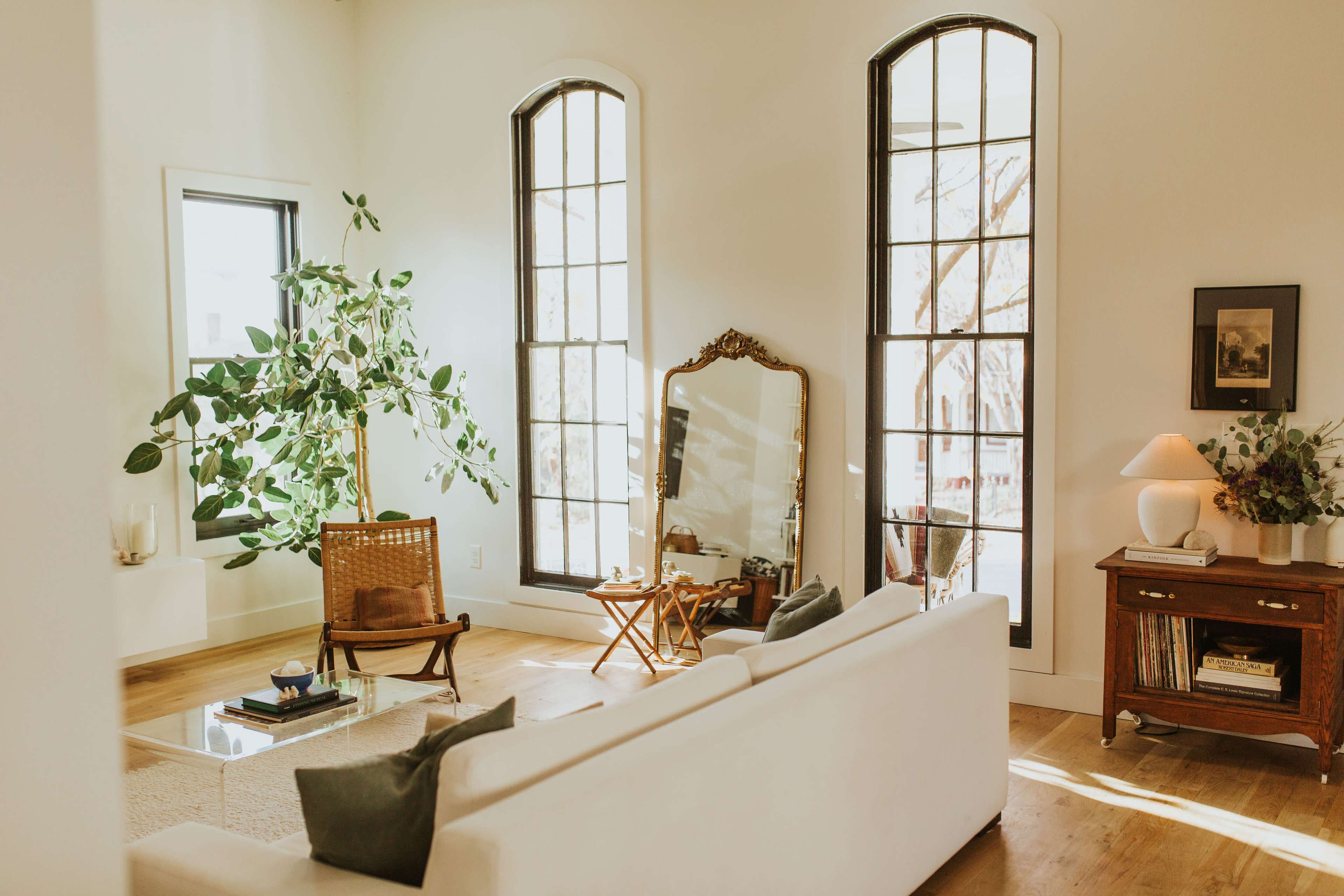 A bright living room features large windows, a green plant, a mirror, and a vintage wooden chair beside a coffee table.