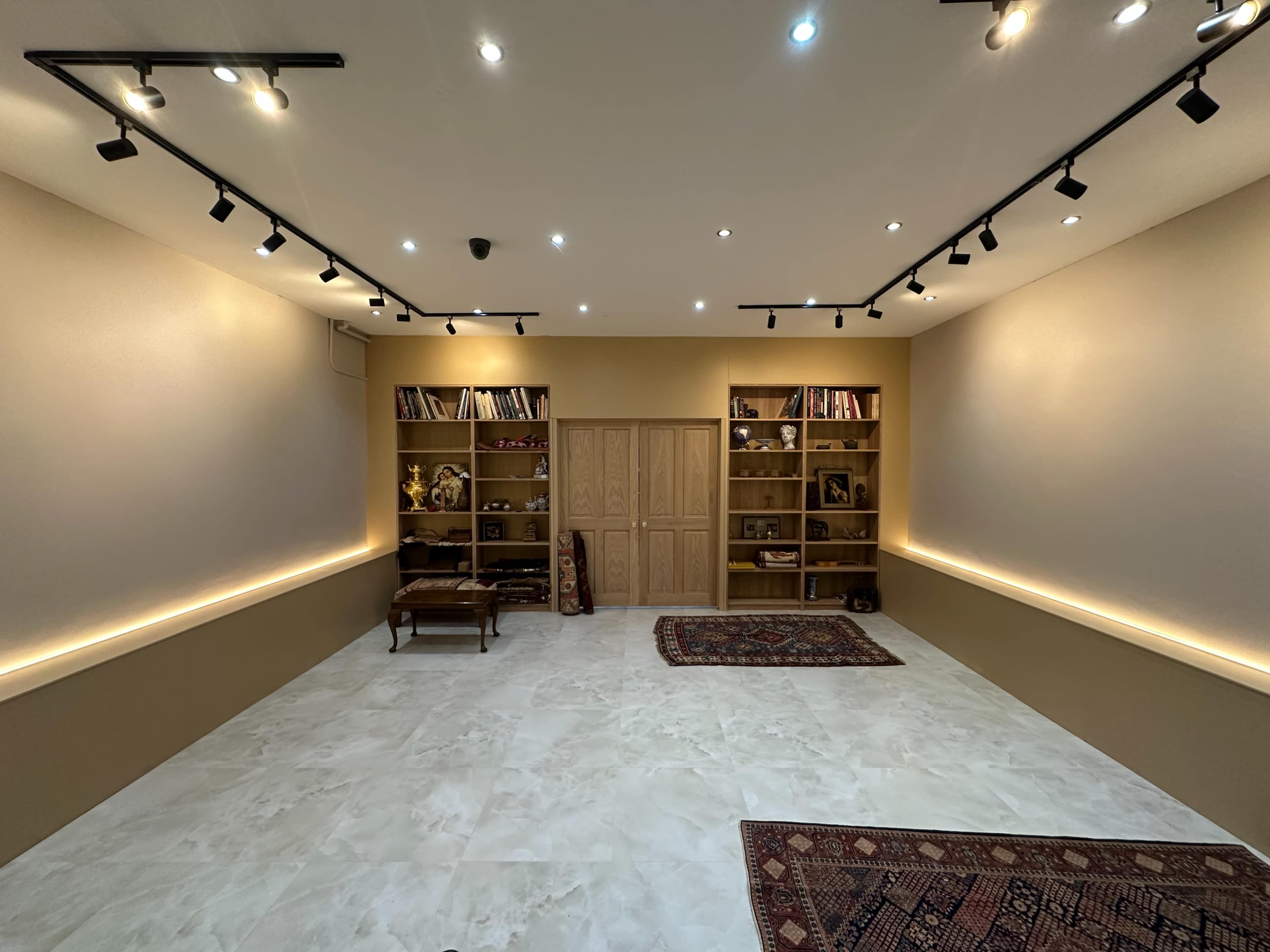The image shows a spacious, well-lit room with shelves filled with decorative items, a wooden cabinet, and a couple of rugs on the floor.