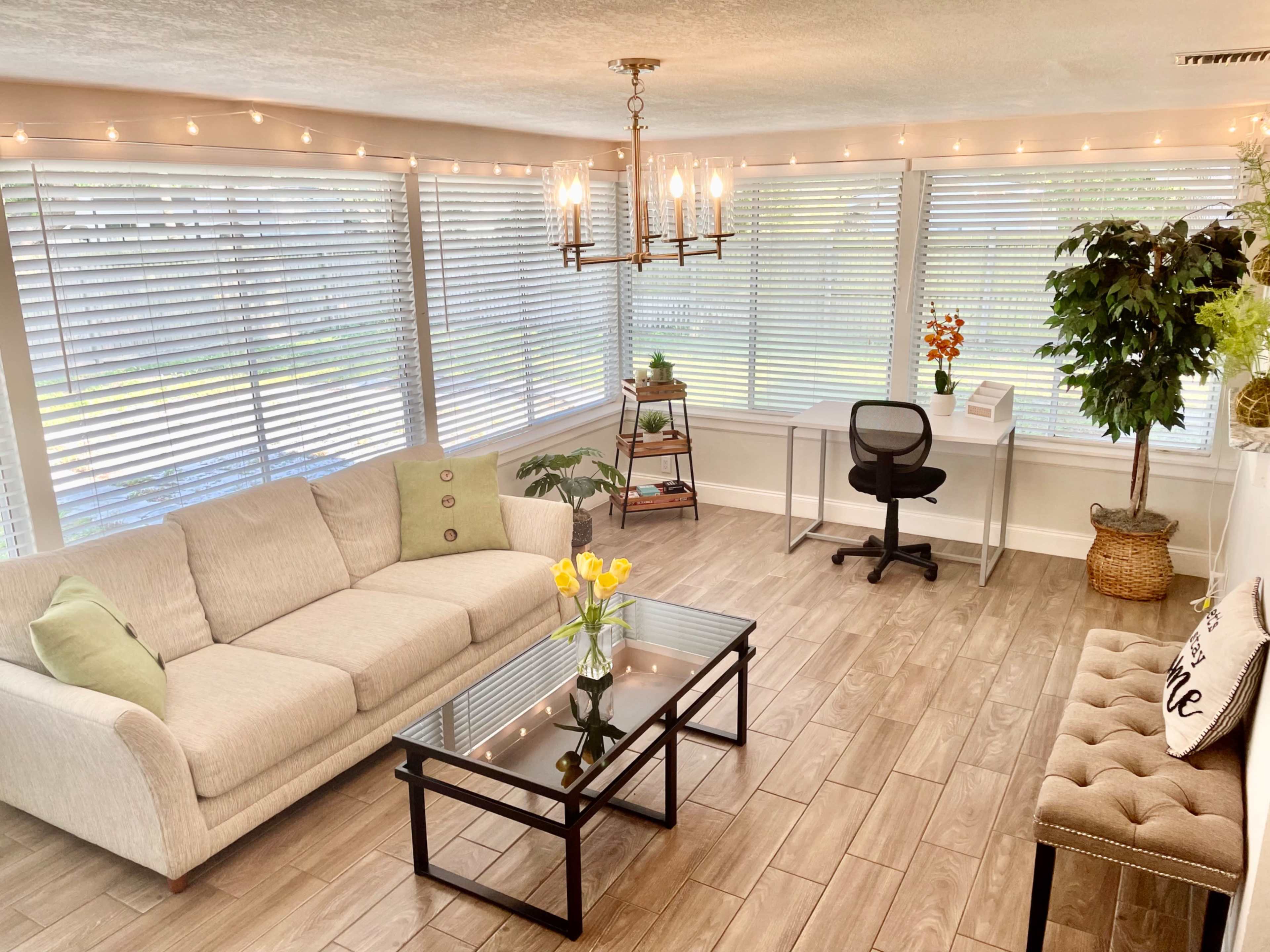 The image shows a well-lit room with large windows covered by blinds, featuring a light-colored sofa, a glass coffee table with a vase of yellow flowers, a black office chair, and a small desk with a decorative plant in the corner.