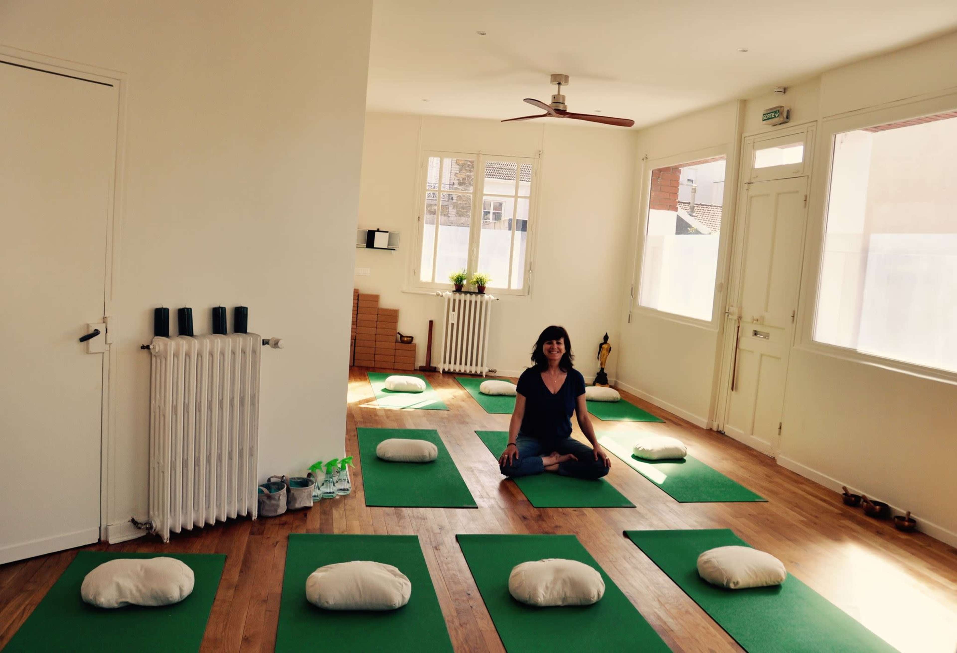 A person sits cross-legged on a green yoga mat in a minimalist studio with additional mats and cushions arranged around her.