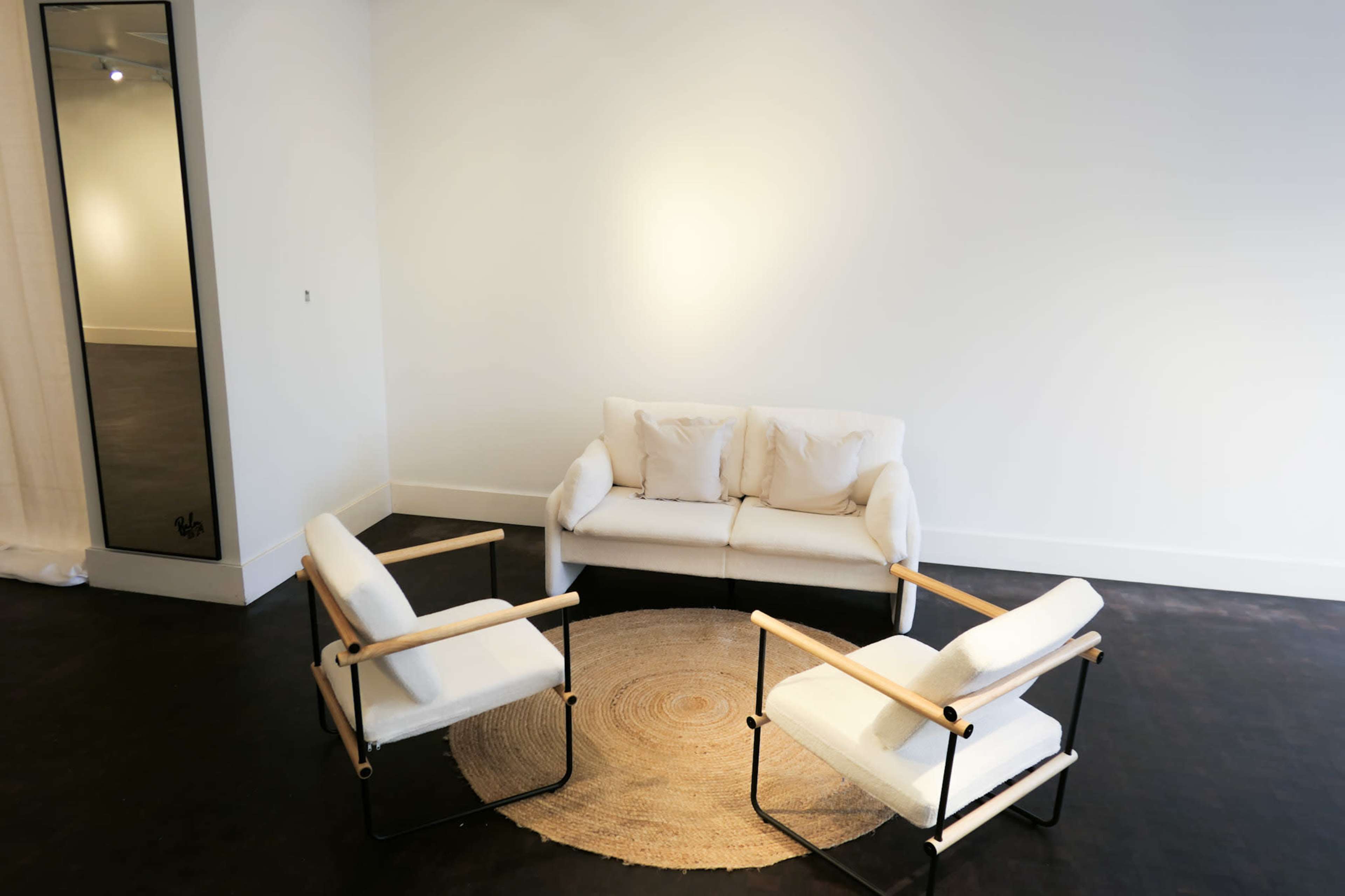A minimalist room features a white sofa and two armchairs arranged around a circular jute rug, with a floor mirror leaning against the wall.
