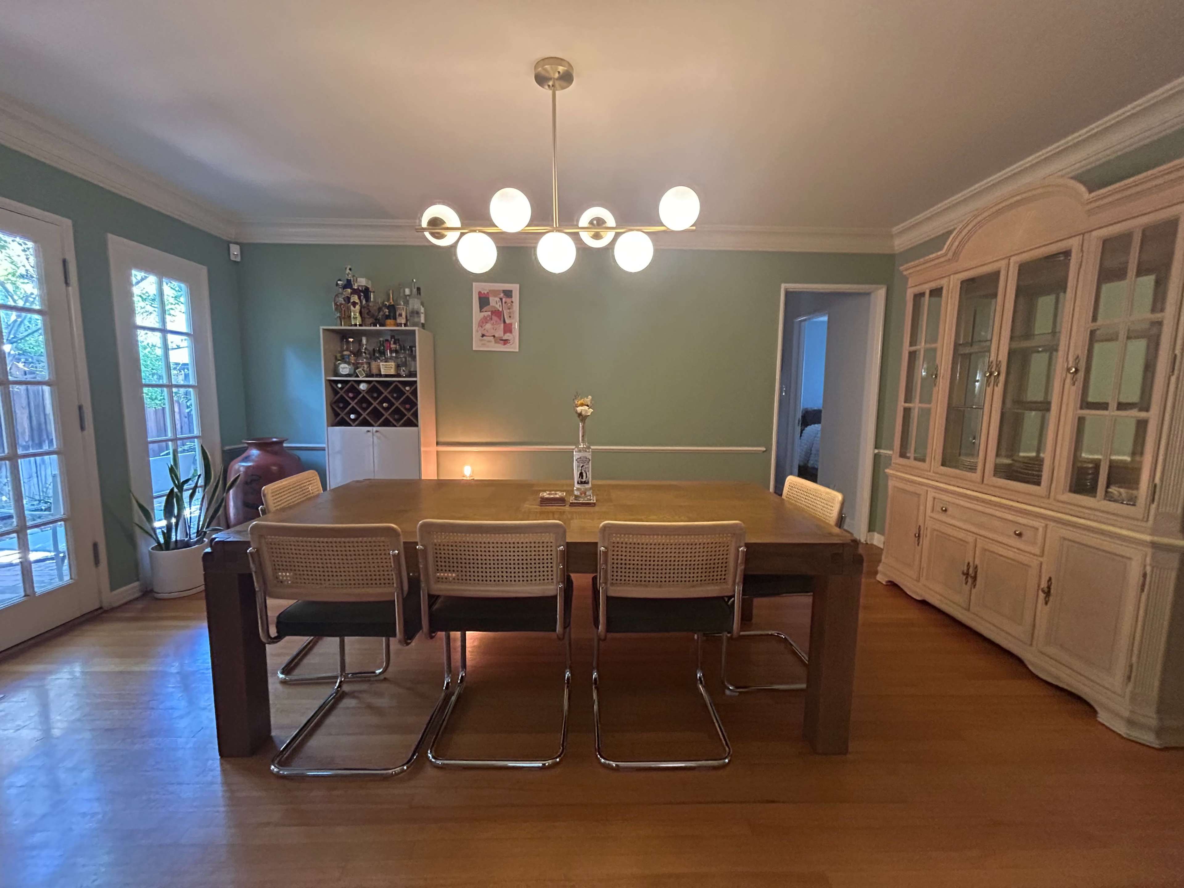 The image shows a dining room with a large wooden table surrounded by four chairs, an open shelving unit, and a cabinet against a green wall, illuminated by a modern chandelier.
