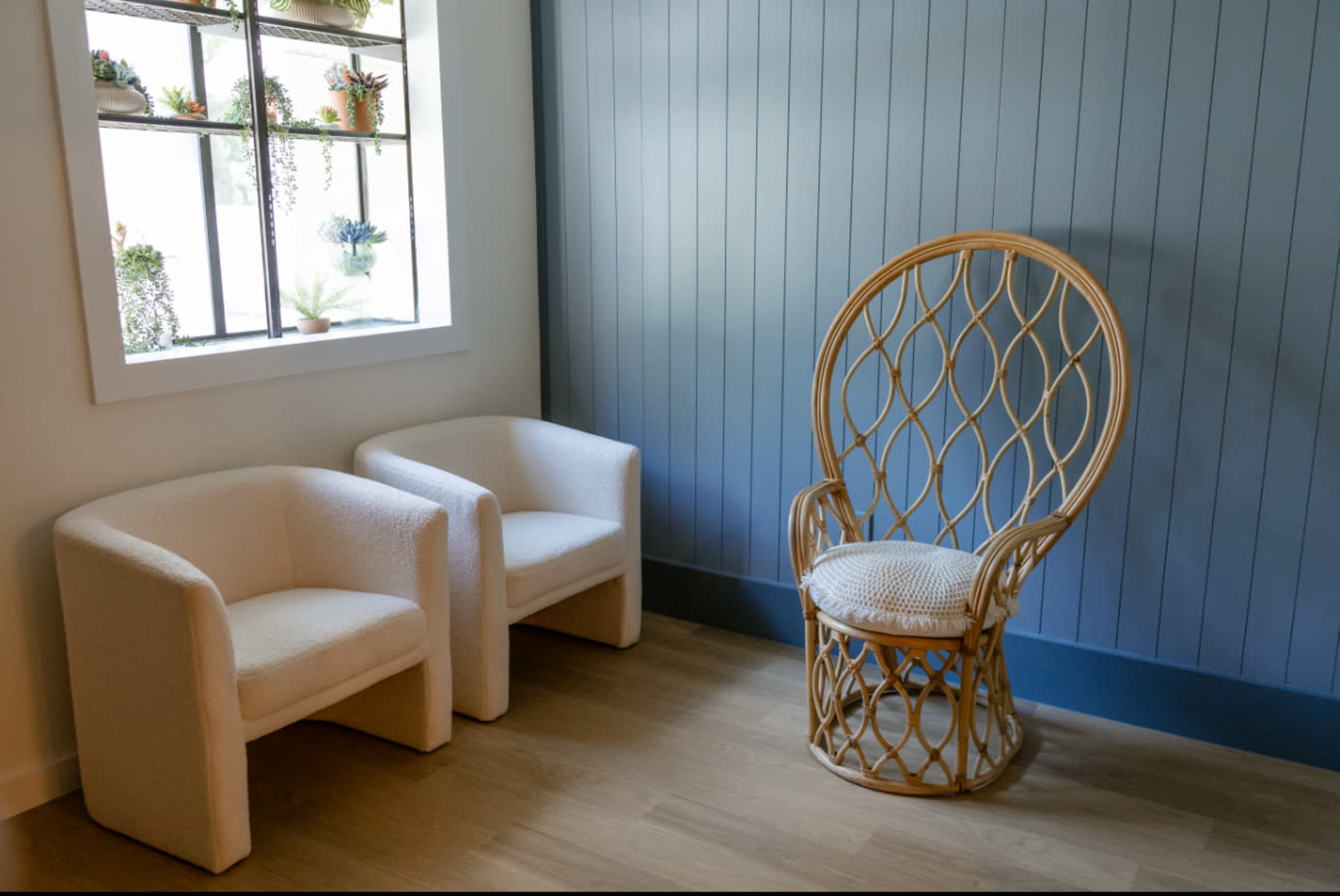 The image shows a cozy corner with two white upholstered chairs and a large wicker chair, positioned next to a window with potted plants.