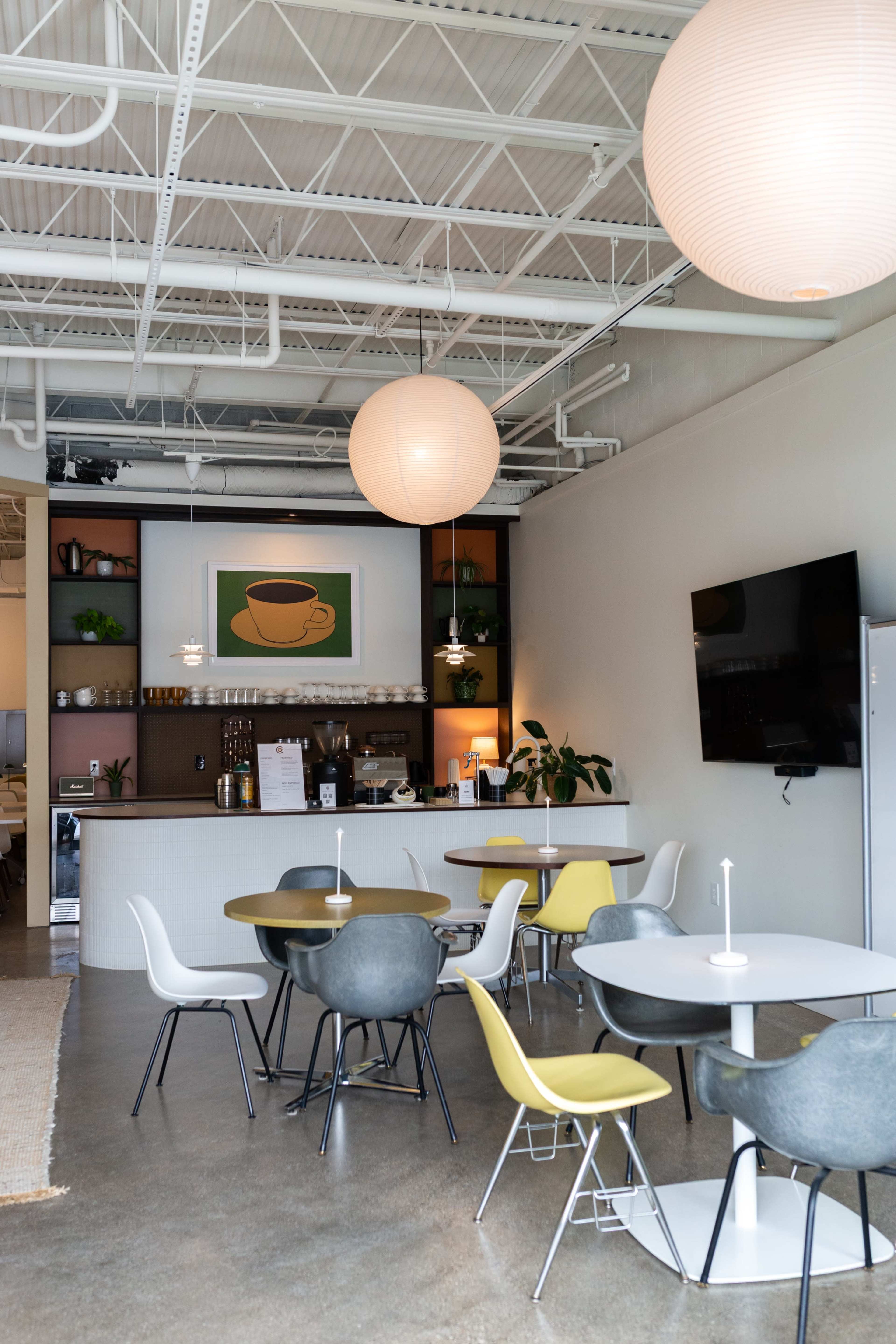 The image shows a modern cafe interior with round tables and chairs, a coffee bar, and large pendant light fixtures.