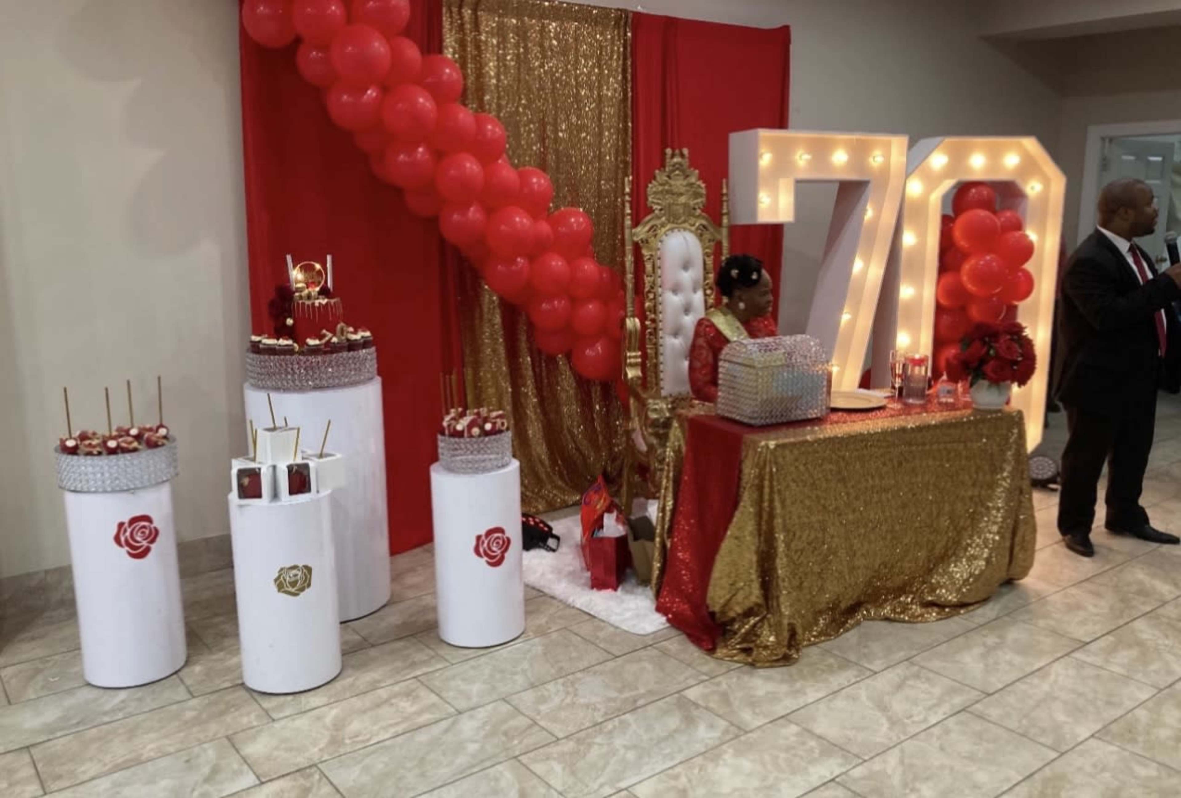 A festive event setup featuring a red and gold theme with a decorated dessert table, large "70" display, and a throne-like chair adorned with balloons.